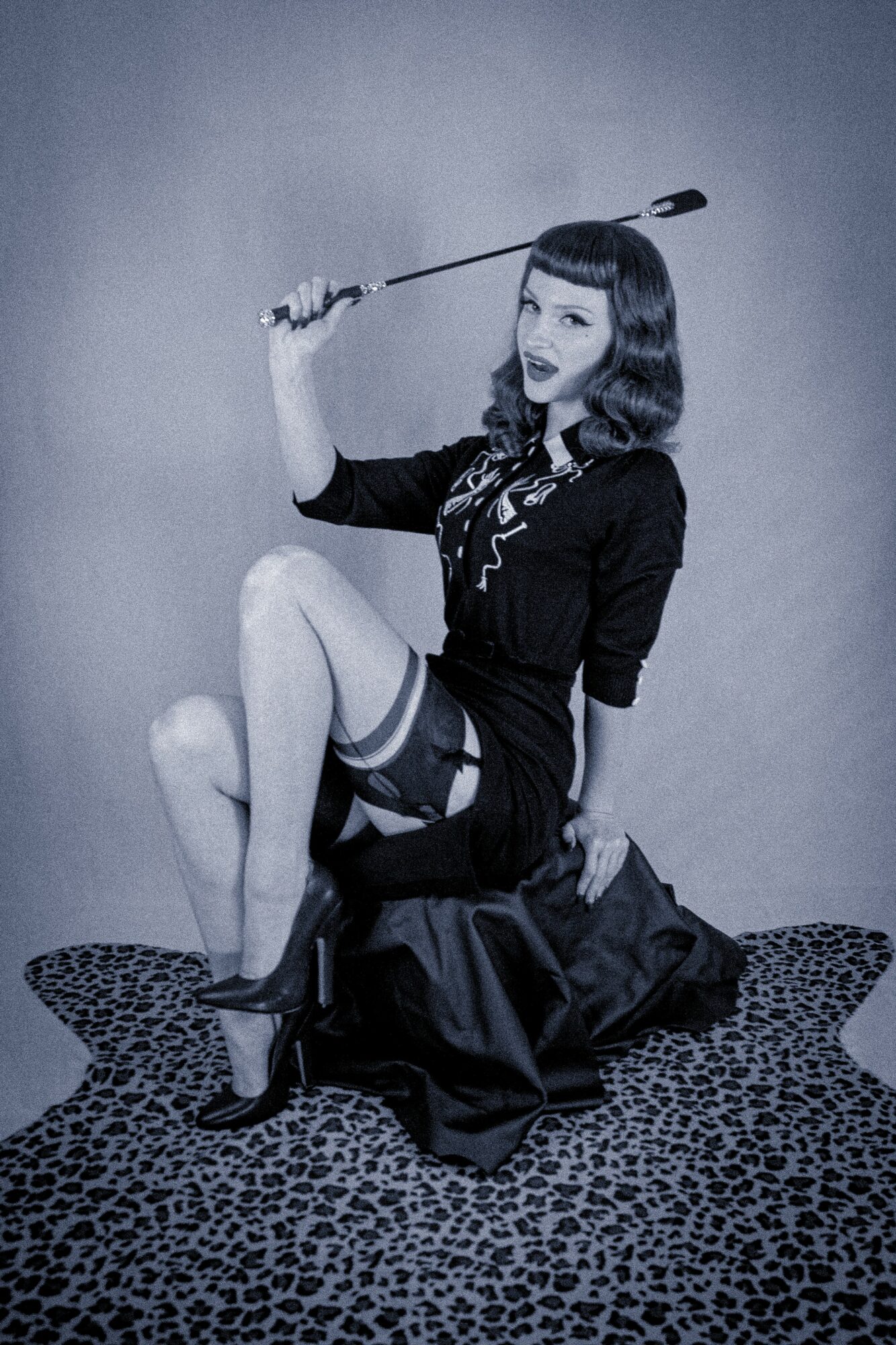 Woman in vintage outfit holding a riding crop, sitting on a leopard-print rug, with a plain background.