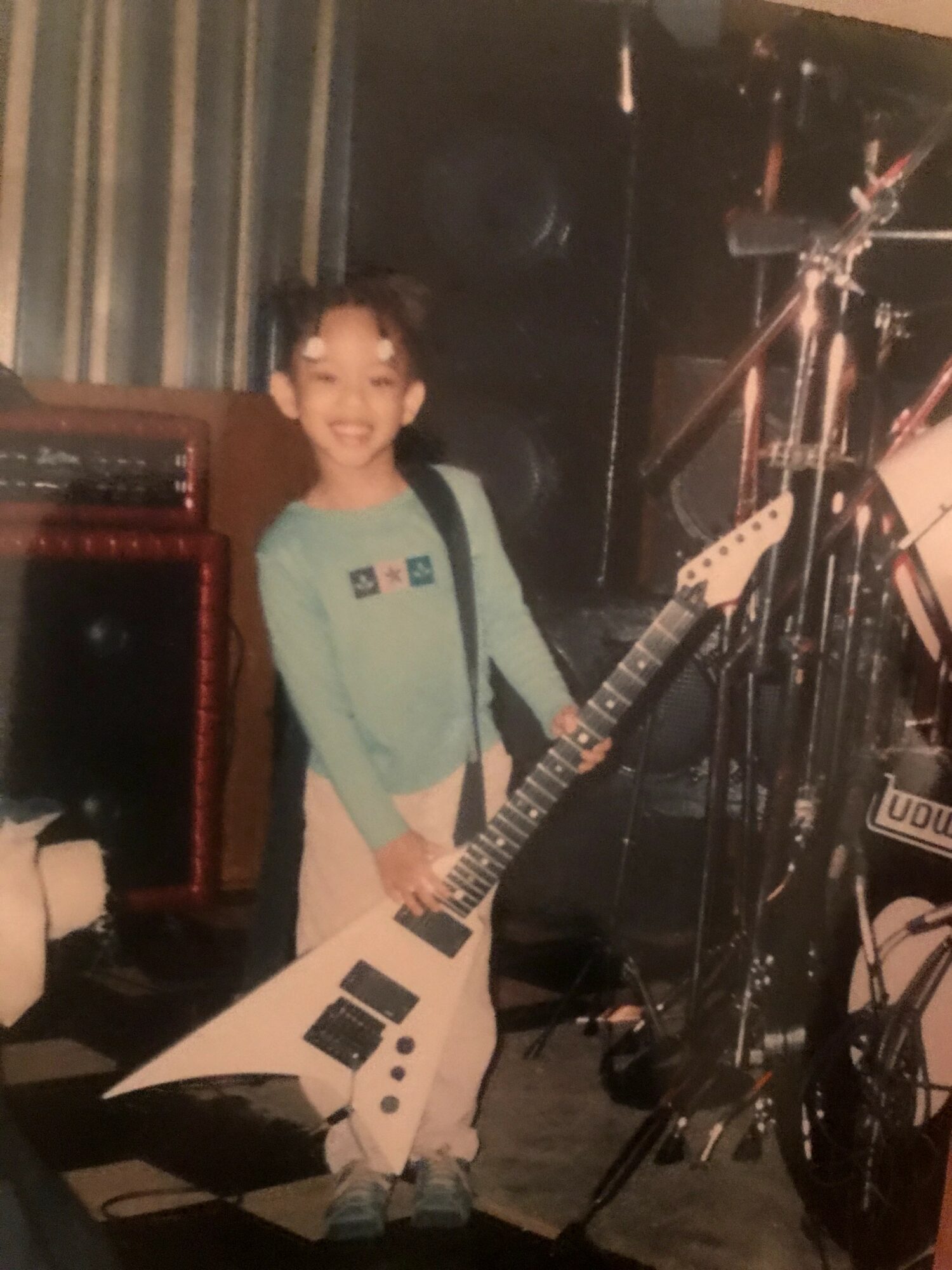 Child holding a white electric guitar on stage, smiling, with musical equipment in background.