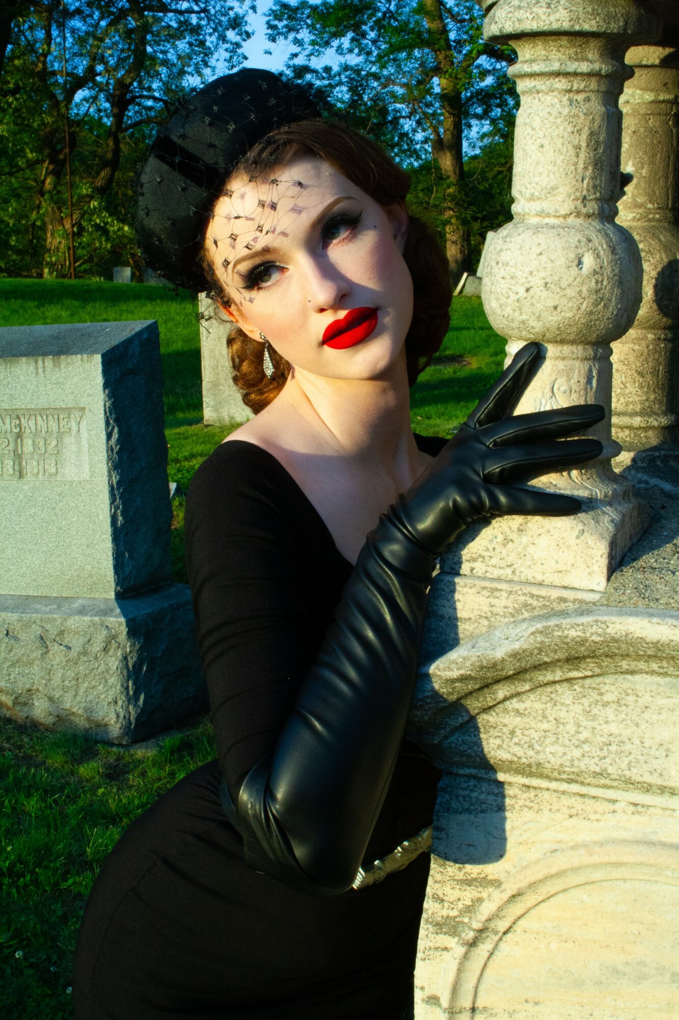 Woman in vintage black dress and gloves outdoors, leaning on stone structure, with trees and blue sky in background.