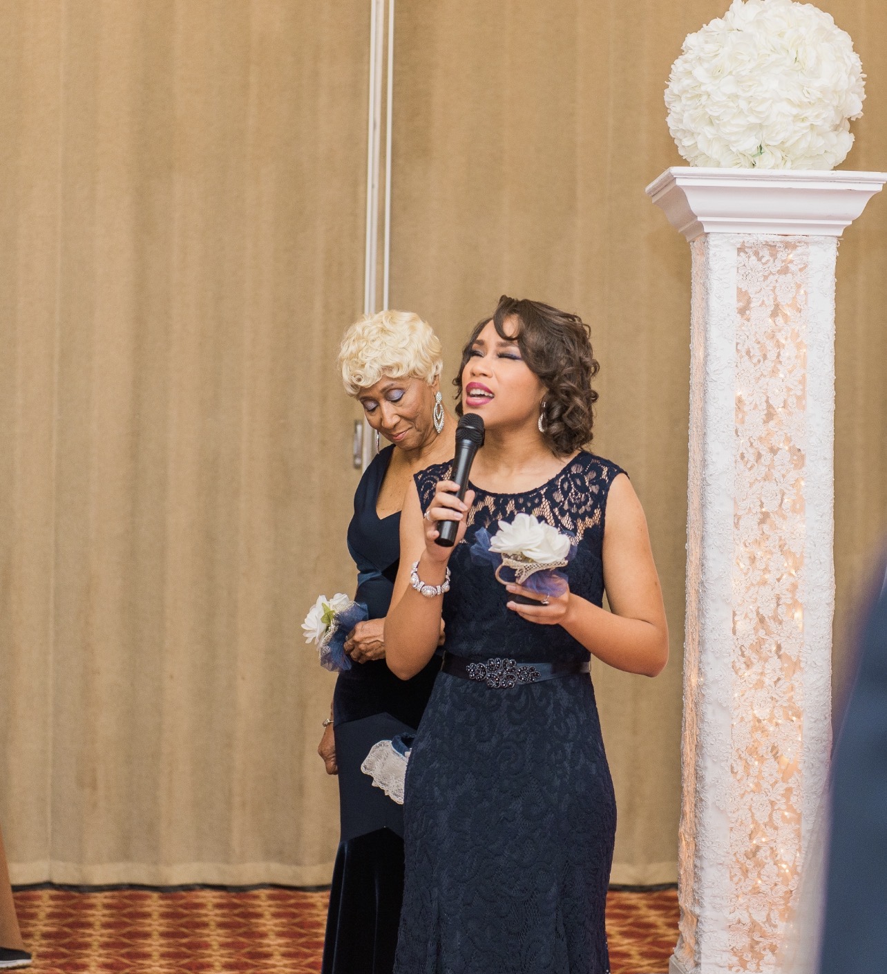 Two women, one singing with a microphone and holding a bowl, the other looking down, in front of a beige curtain and floral column.
