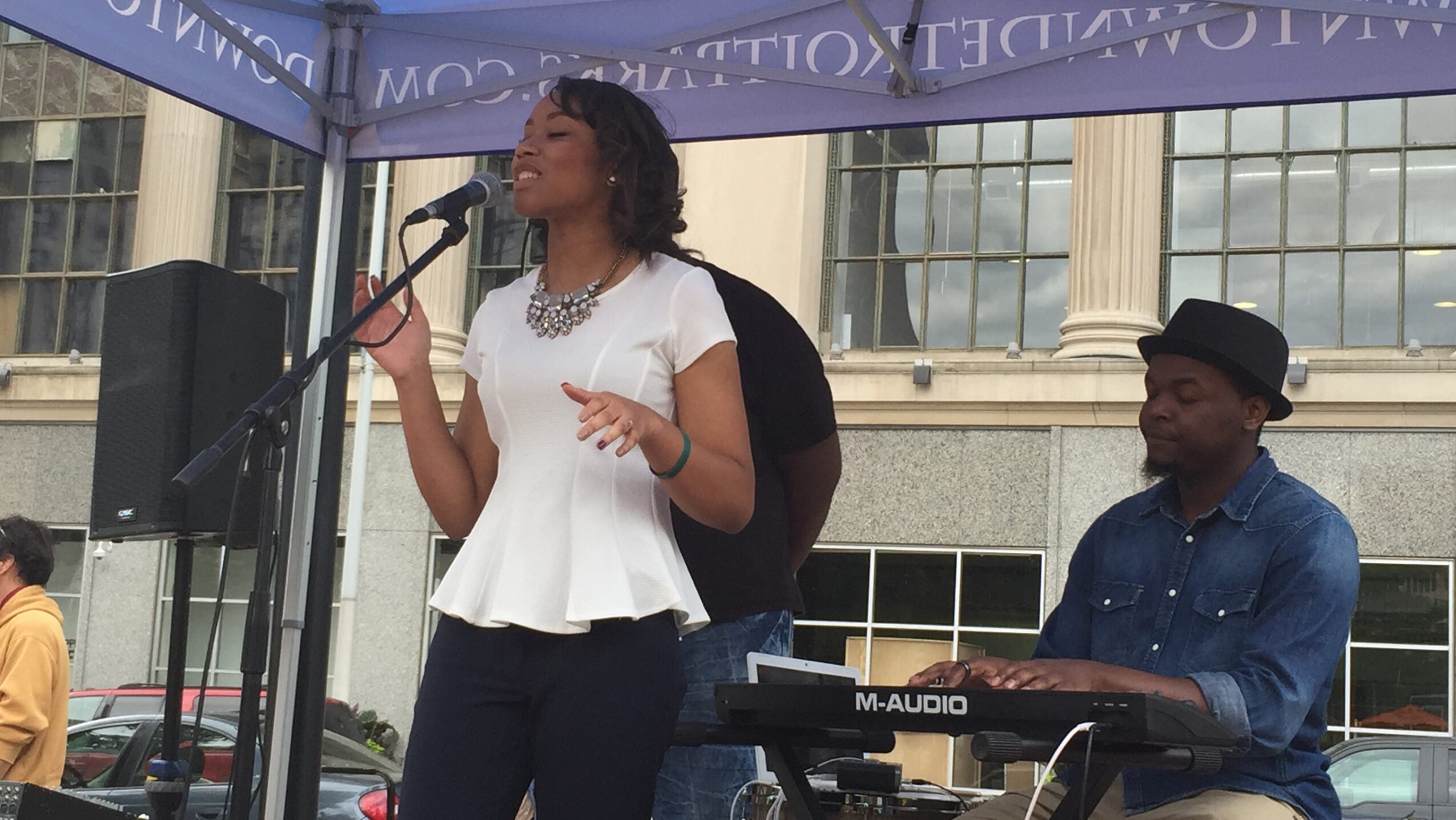 A woman singing into a microphone on stage with a keyboard player beside her, outdoors in front of a building.