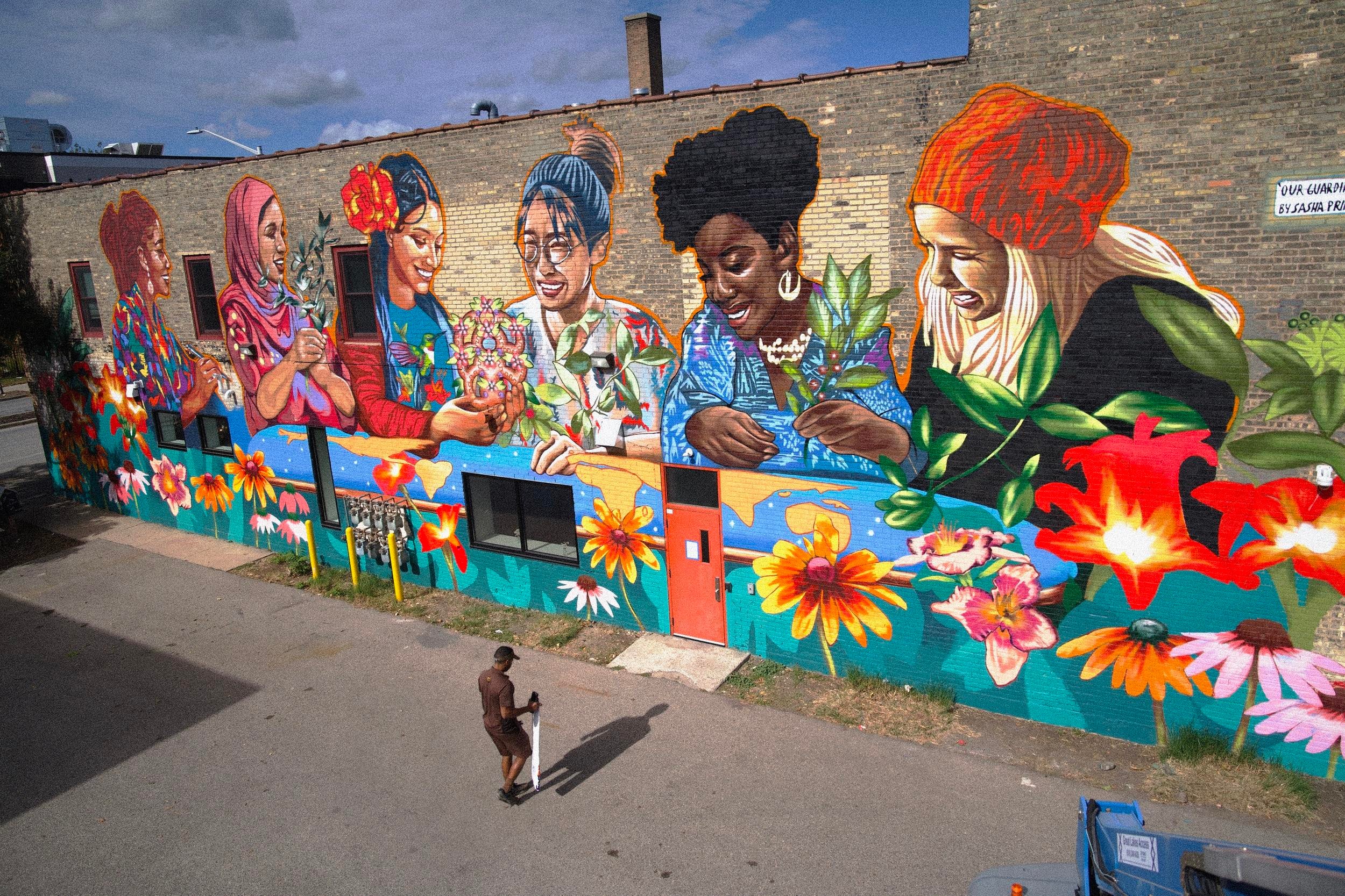 Colorful mural of diverse women with flowers, painted on a brick wall, with a person walking nearby. Word 'LOVE' visible.