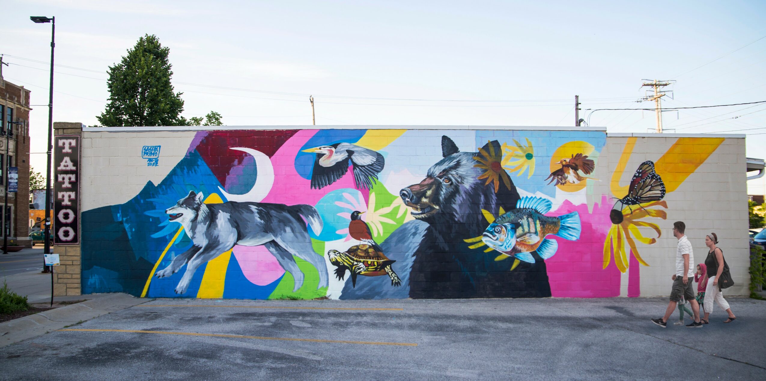 Colorful mural of cats with butterflies and flowers on a wall, with people walking nearby.