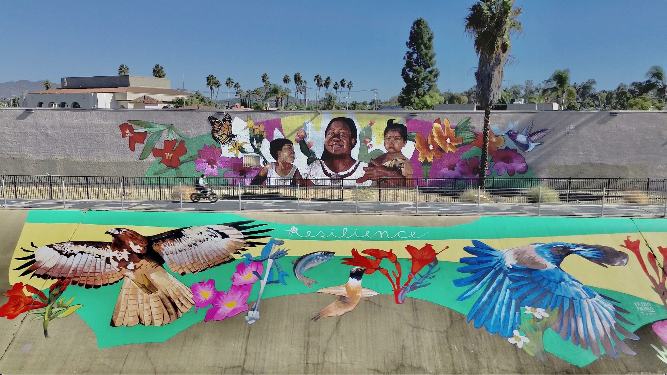 Colorful mural of children and birds painted on a wall and a bridge, with a cityscape and trees in the background.