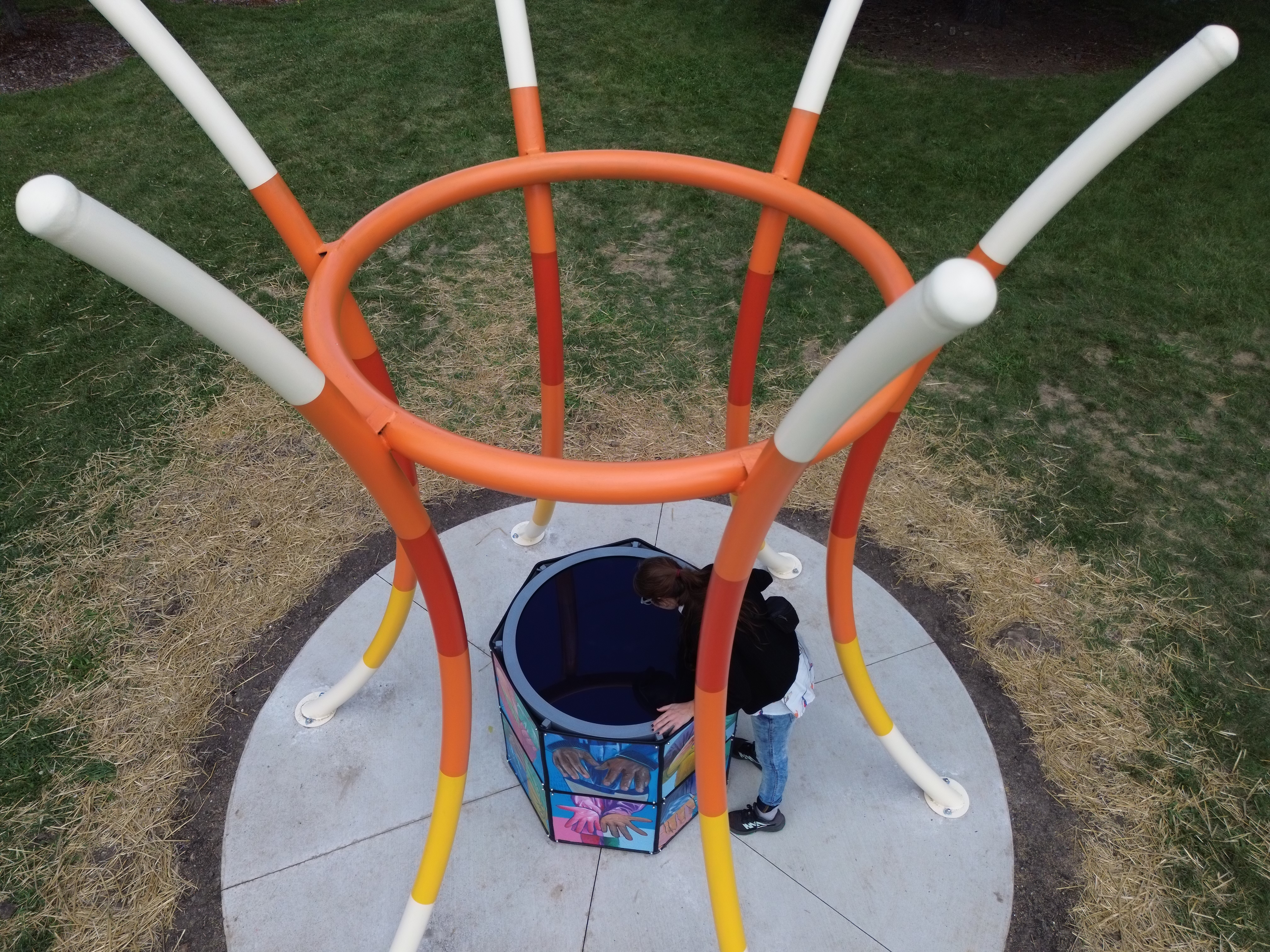 Child playing in a colorful bucket under a circular playground structure with orange, white, and yellow supports on grass.