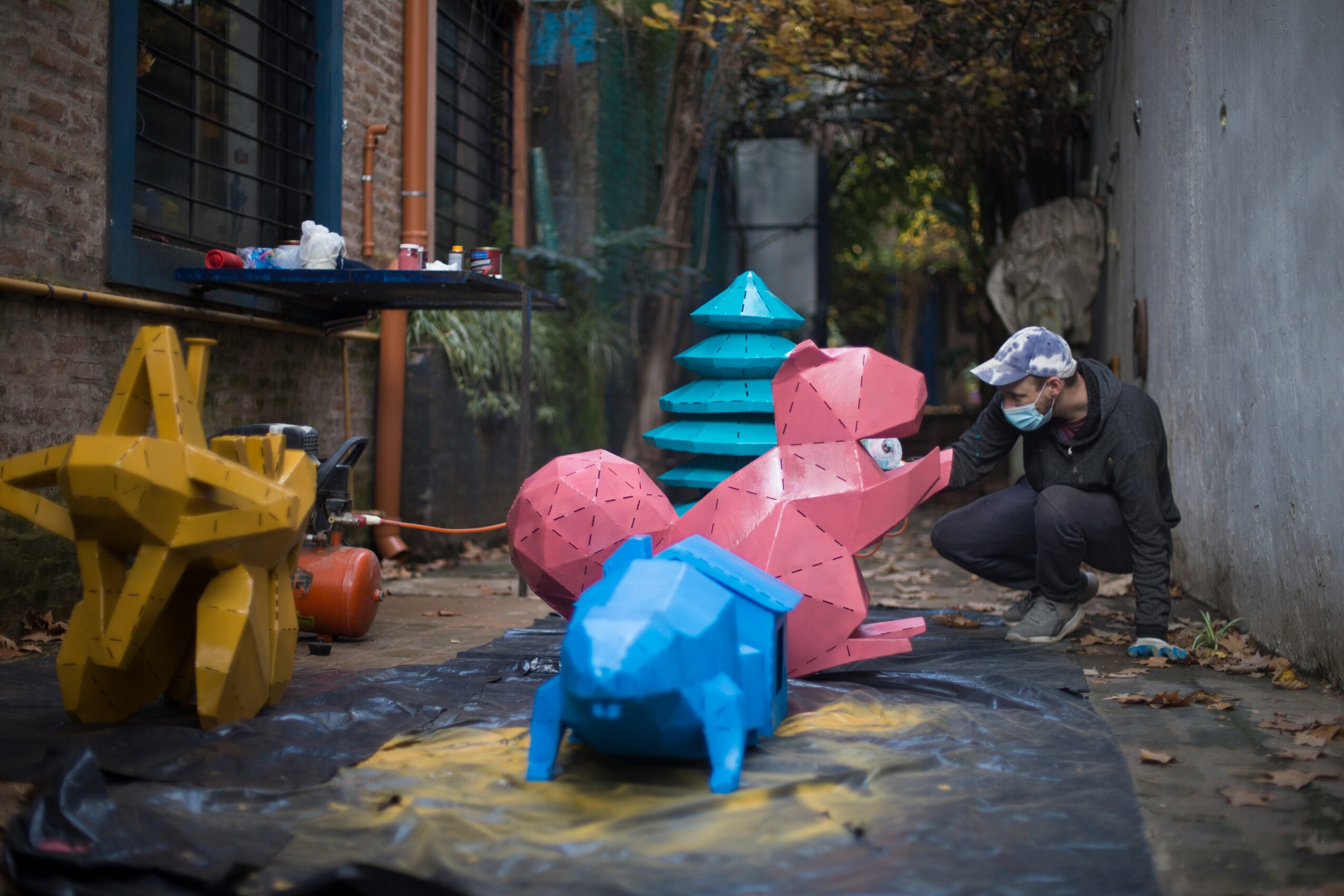 Person crouching near colorful animal sculptures outdoors, with trees and a building in the background.