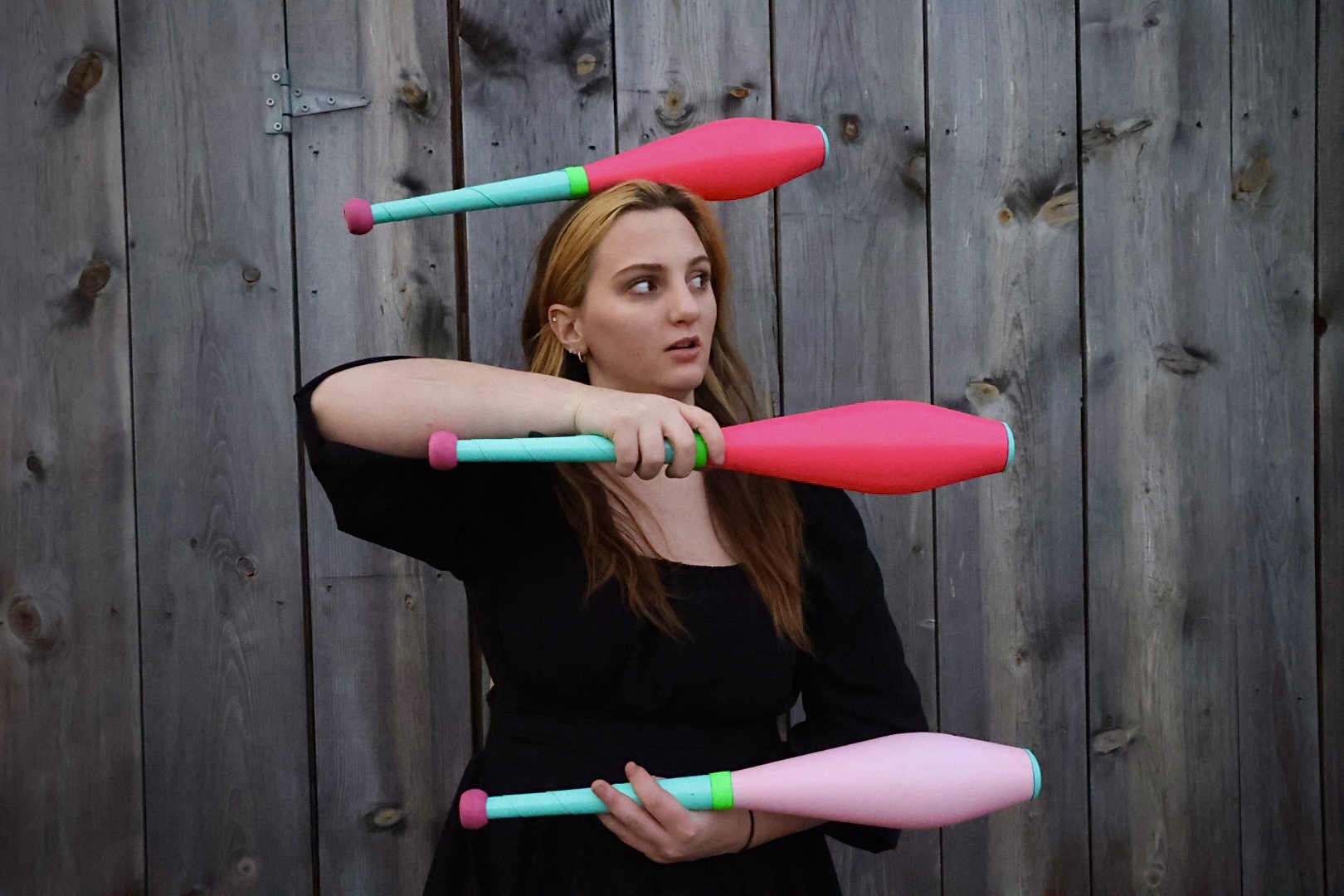 Young woman with long hair holds juggling clubs in front of a wooden fence, balancing a club on her head.