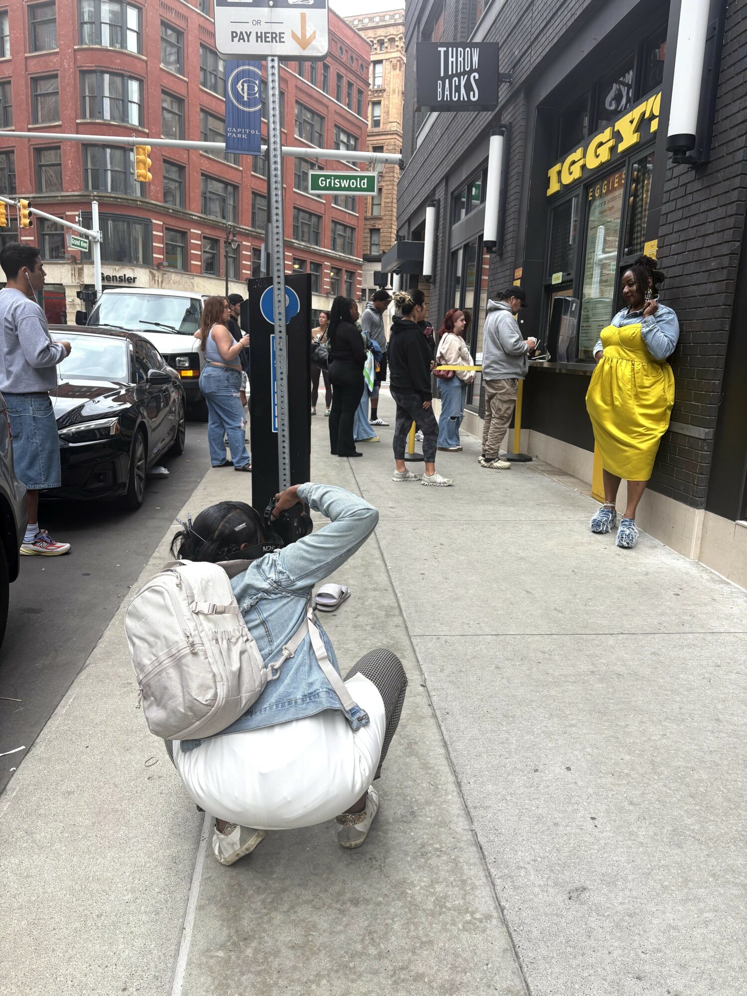Person crouching on sidewalk with backpack, surrounded by pedestrians and buildings, some with signs and storefronts.