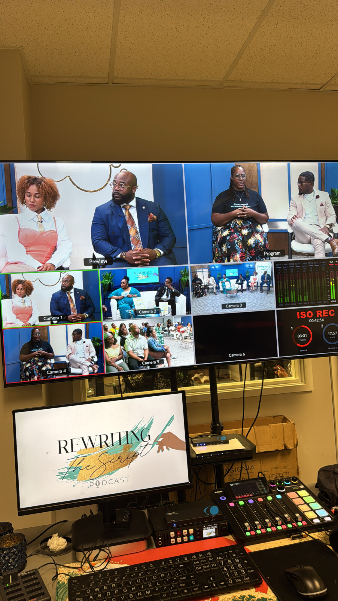 Multiple people seated and standing in a panel discussion, with a screen displaying a presentation and a control panel below.