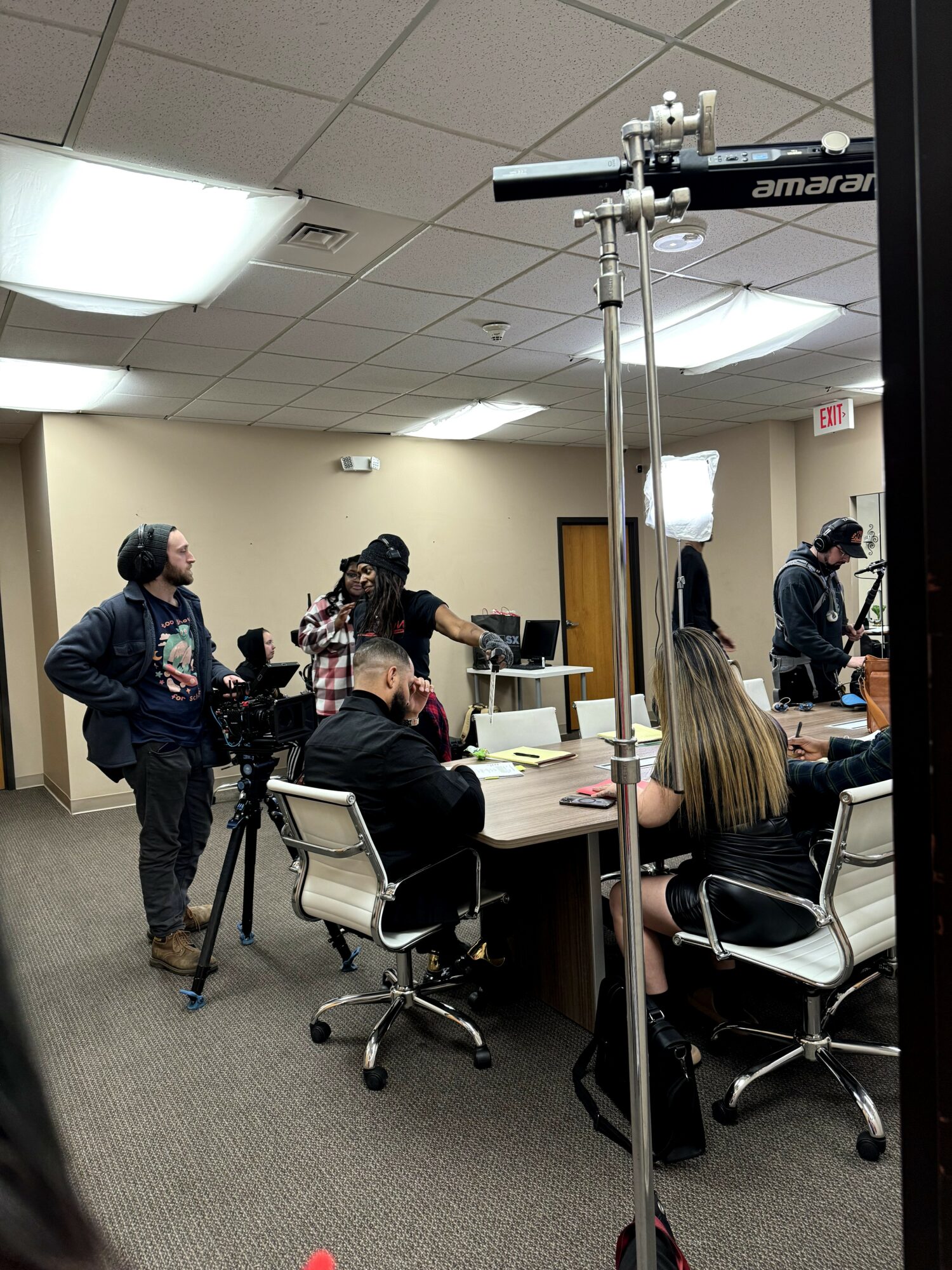 People in a conference room with cameras and equipment, some seated at a table, others standing and talking.