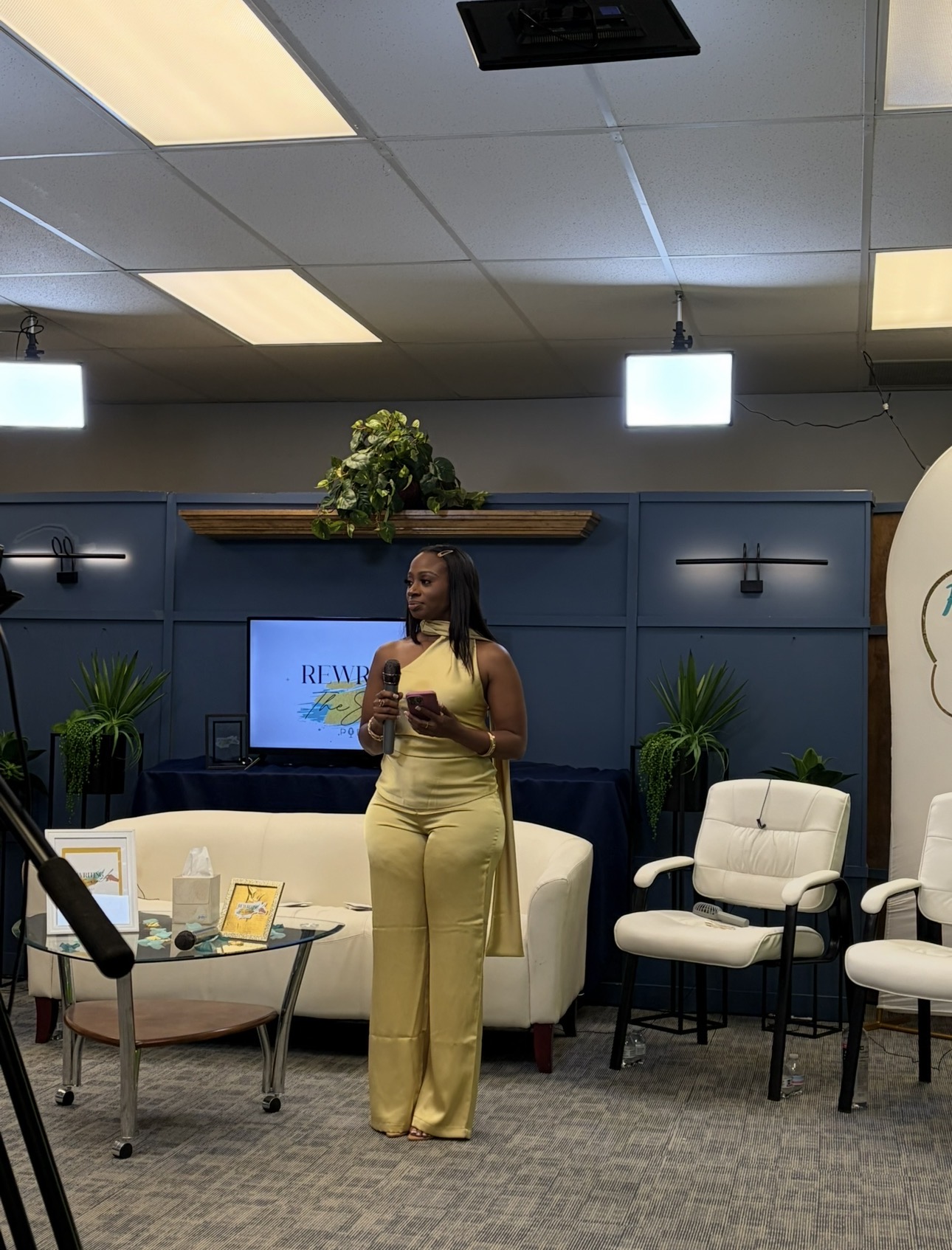 Woman in yellow pants standing in a room with blue walls, white chairs, and a television.