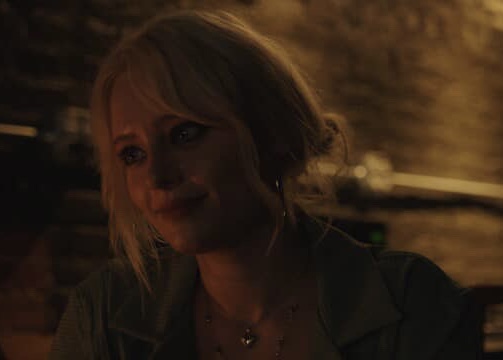 Woman with blonde hair in a dimly lit setting, smiling slightly, with a brick wall background.