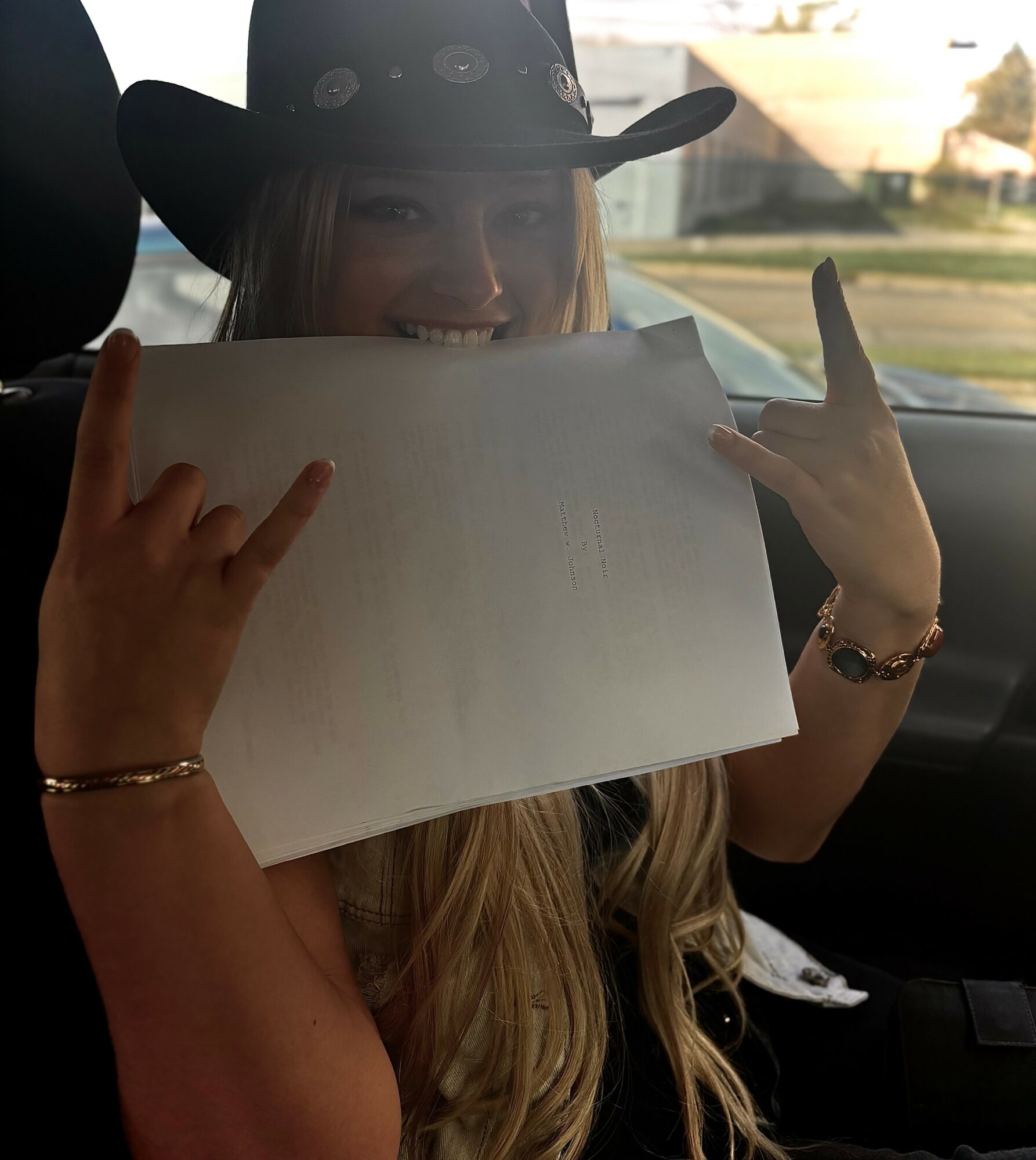 Young woman wearing a cowboy hat holding a paper, smiling, making rock signs with both hands, inside a vehicle.