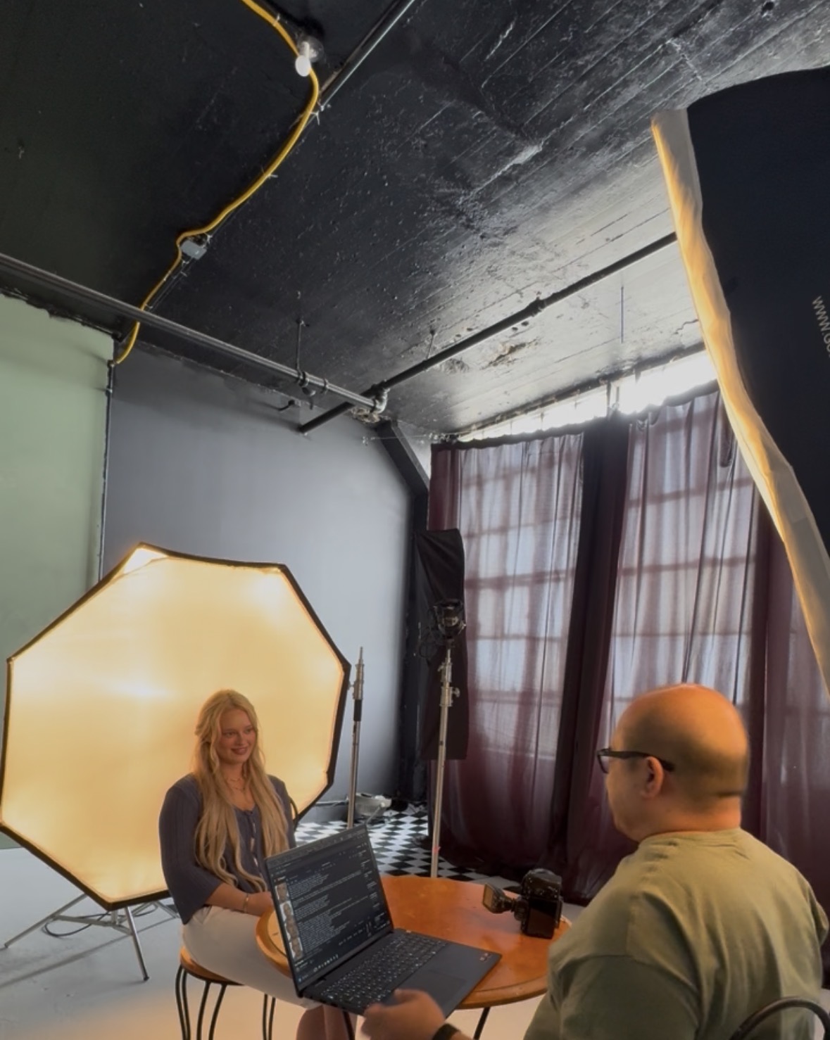 Two people in a photography studio with lighting equipment and curtains, one using a laptop, the other sitting nearby.