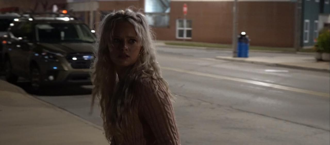 Person with long hair standing on sidewalk at night, street and parked cars in background.