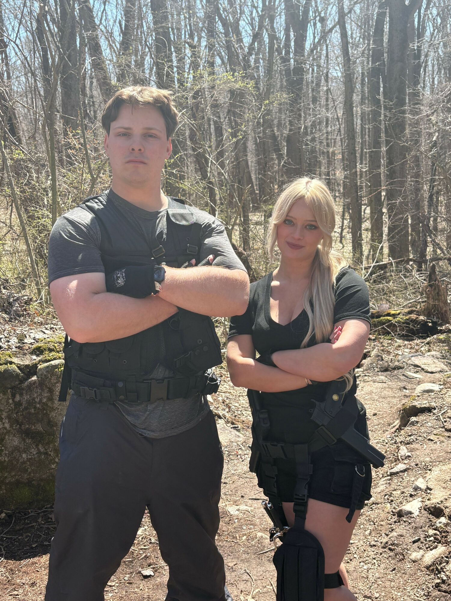 Two people standing outdoors on a dirt path with trees in the background, one with arms crossed and the other with hands on hips.