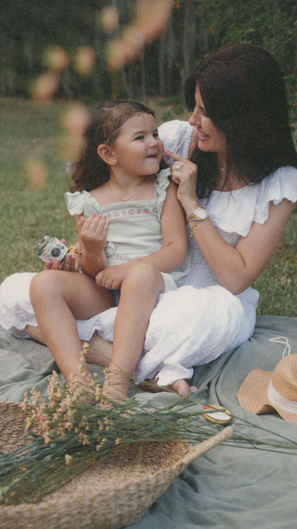 Woman and young girl sitting outdoors on grass, smiling and looking at each other, surrounded by greenery and flowers.