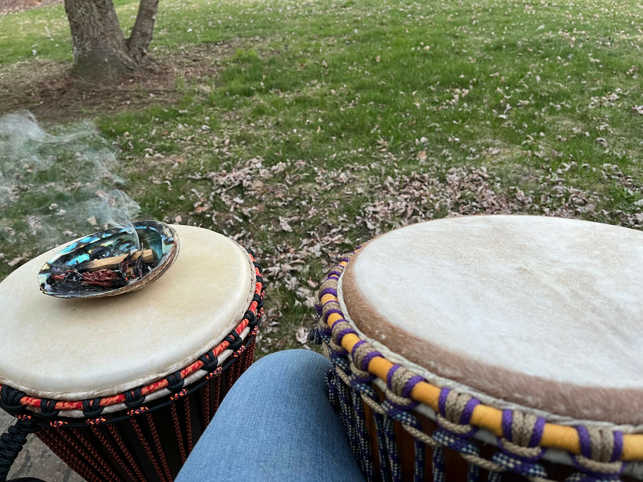 Two drums with drumsticks outdoors on grass, one with smoke rising, and a person's knee visible.