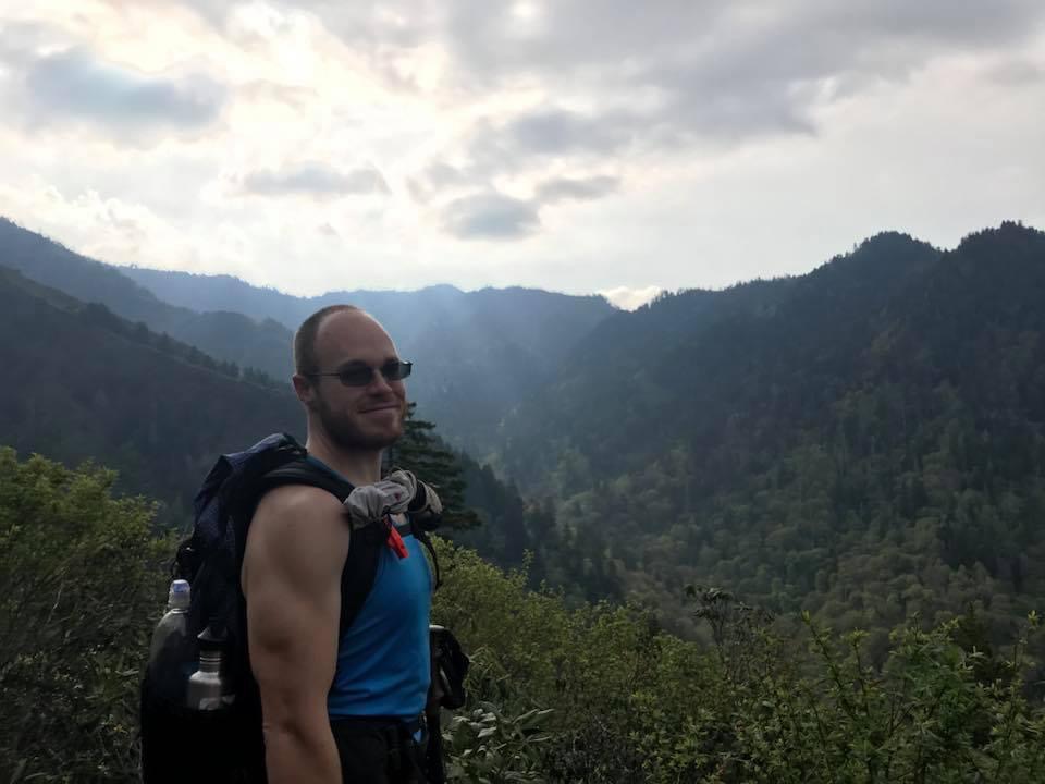 Man with sunglasses and backpack standing outdoors with mountains and cloudy sky in background.