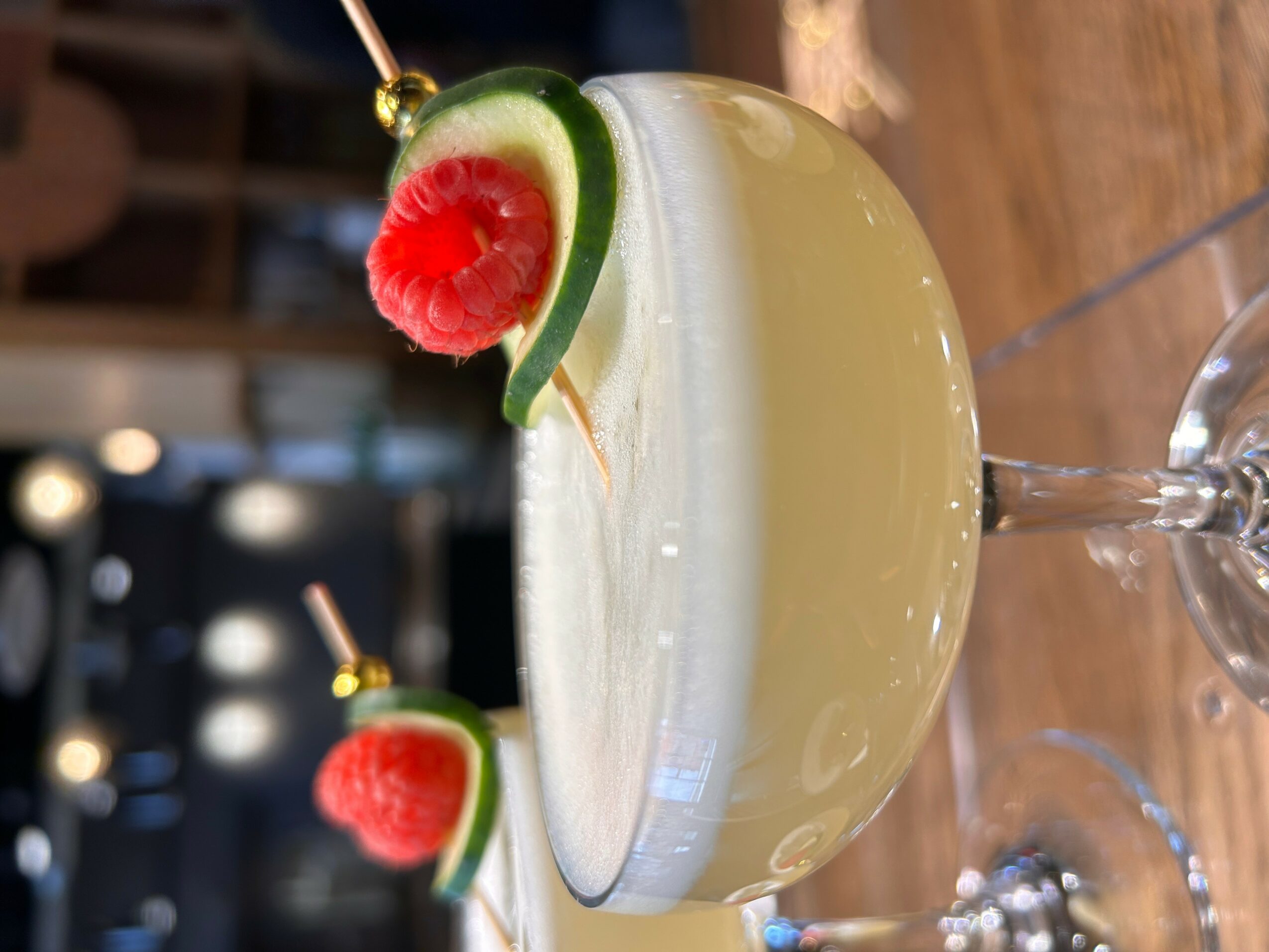 Cocktail glass with a yellow drink, garnished with a lime slice and raspberry, on a wooden surface.
