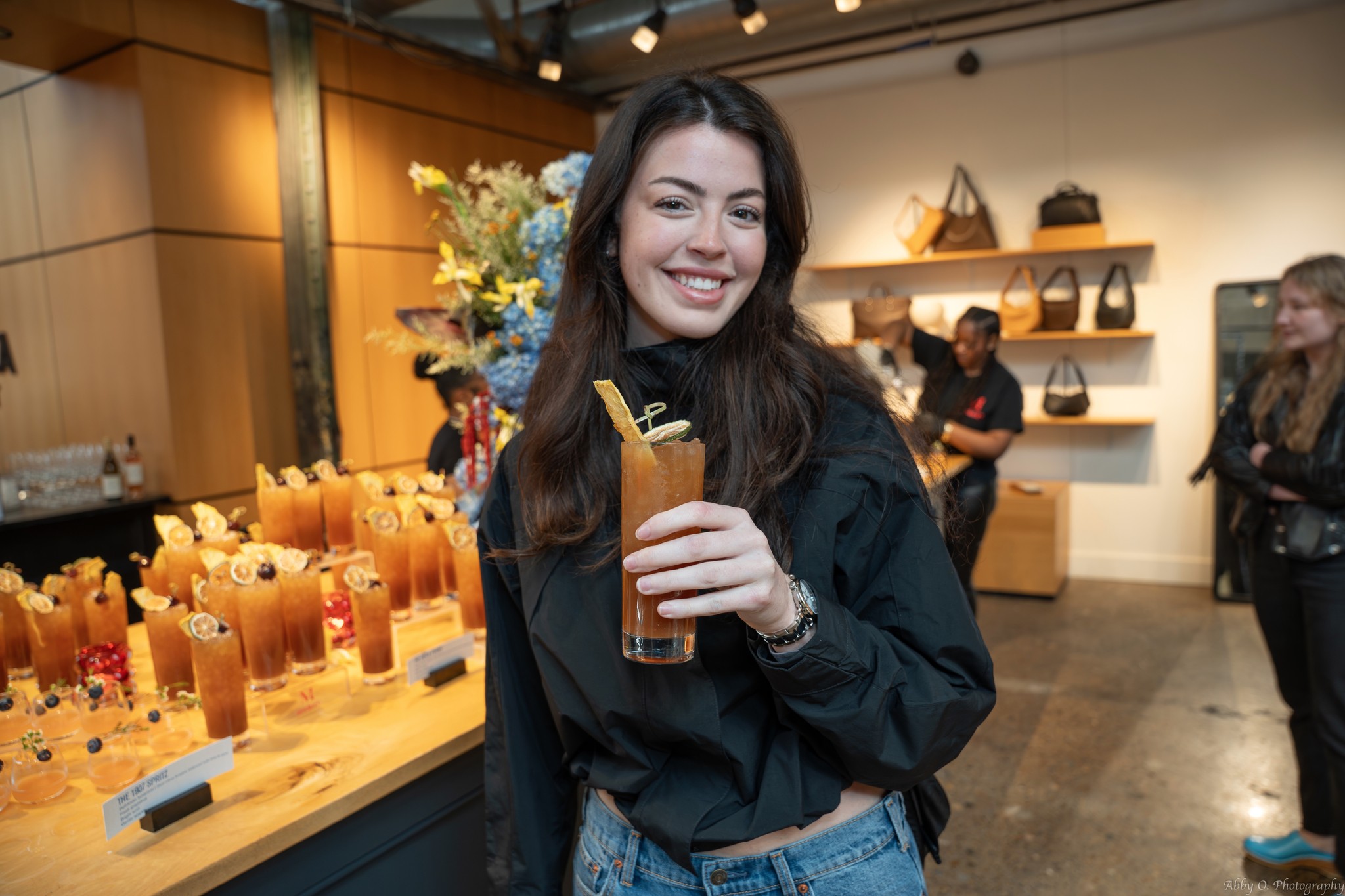 Smiling woman holding a drink in a cafe or bar with a wooden counter and flowers in the background.