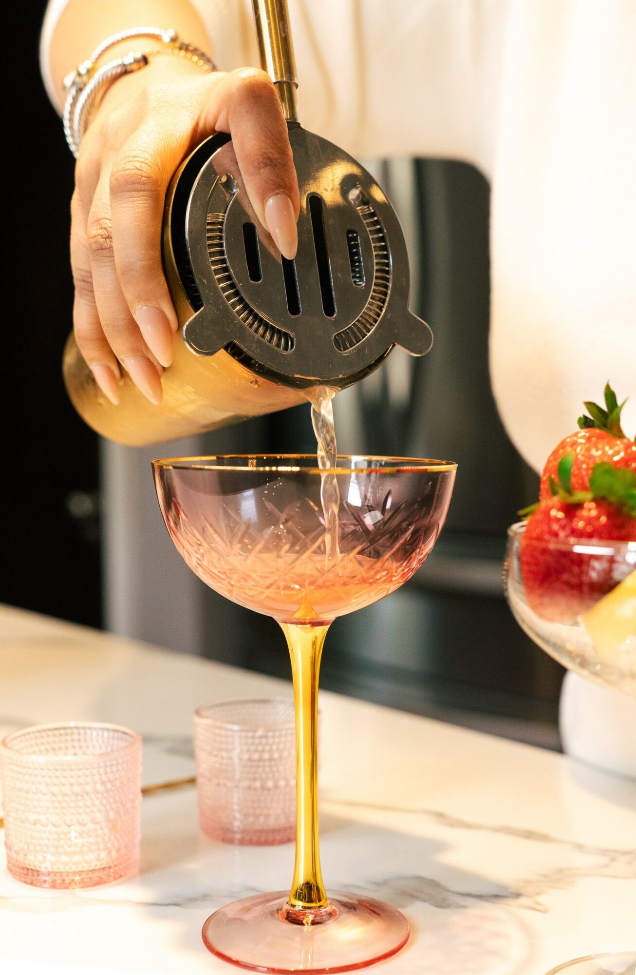 Person pouring a drink into a pink cocktail glass with strawberries nearby, on a white surface.