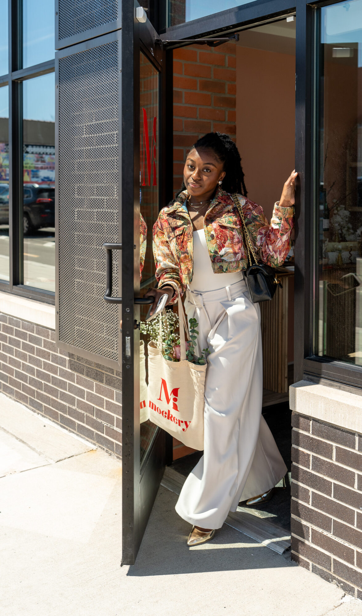 Woman standing in doorway holding shopping bag outside building with large windows and brick wall.