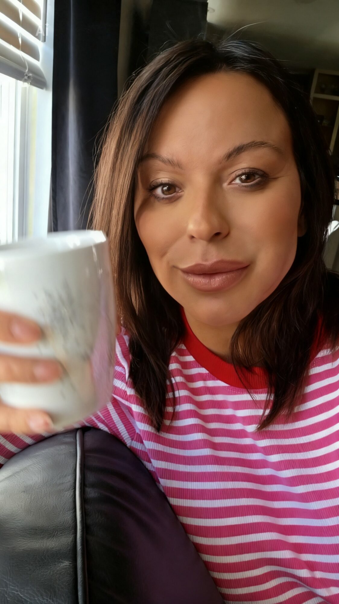 Woman with shoulder-length dark hair holding a cup, wearing a pink and white striped shirt, sitting indoors near a window.