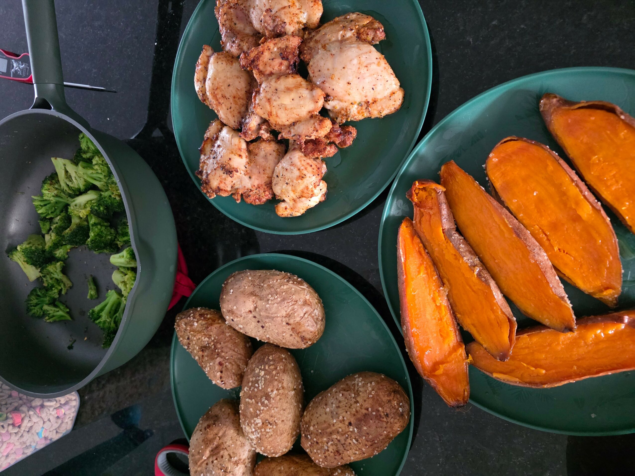 Three bowls of cooked food: broccoli, fried chicken pieces, breaded potatoes, and roasted sweet potatoes on a black surface.
