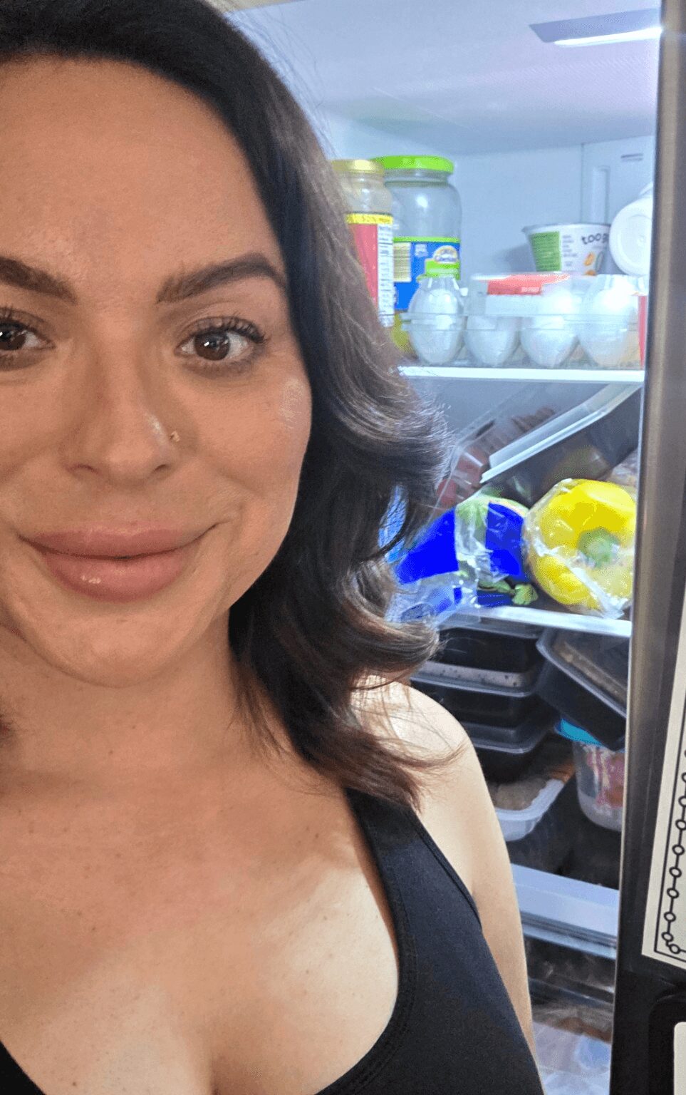 Close-up of a woman with dark hair and a black top, smiling, in front of an open refrigerator with food items.