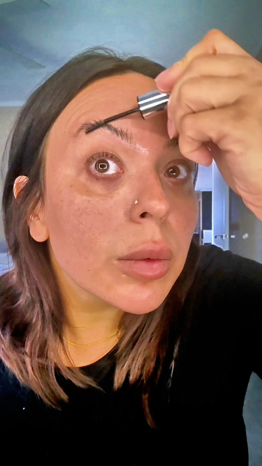 Woman applying mascara to her eyelashes, looking into a mirror, with a focused expression, dark hair, and a nose piercing.