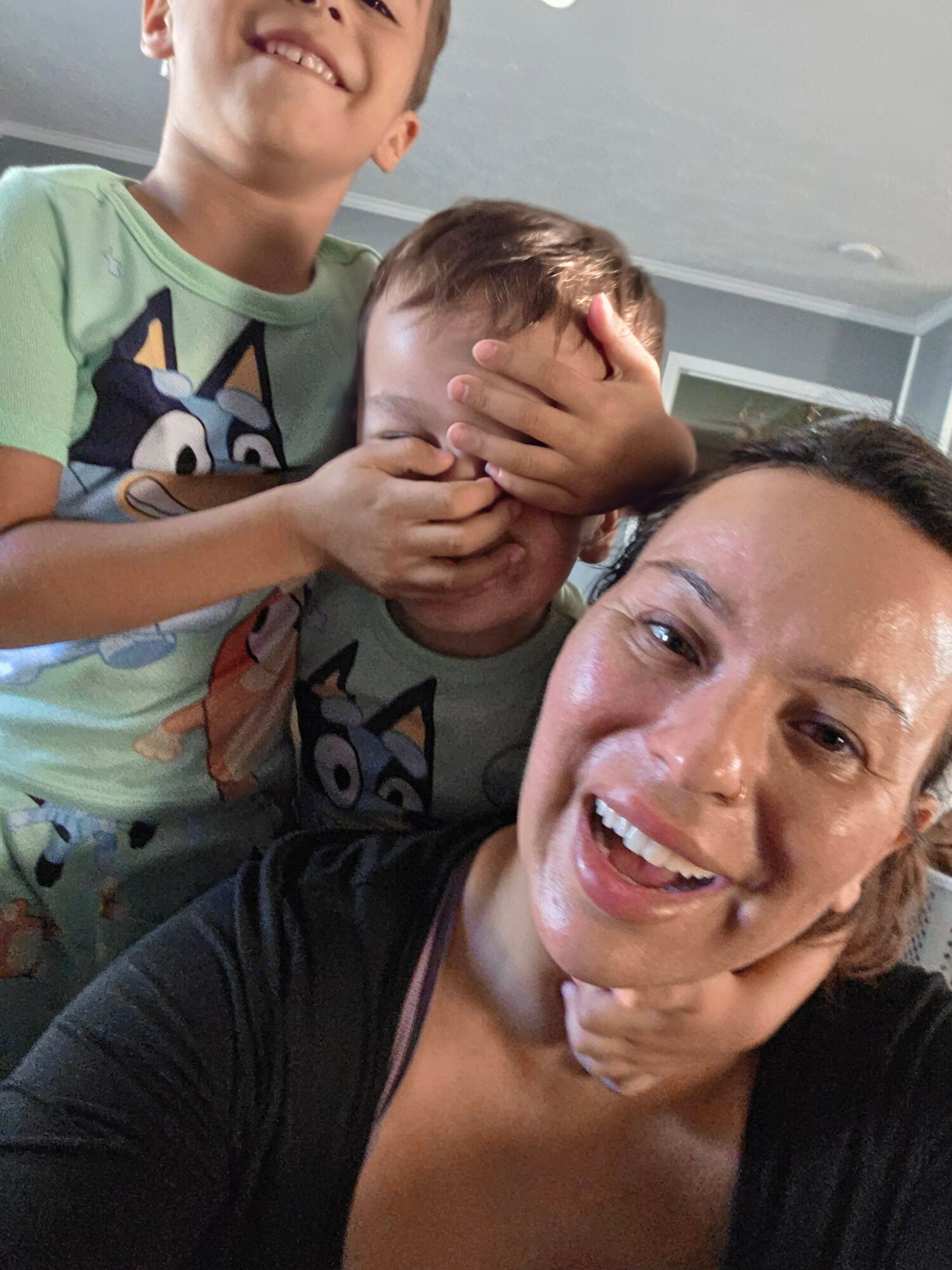 Two children and an adult smiling, with one child covering their face, in a room with a bed and window.