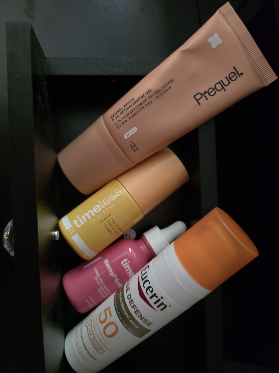 Four skincare products, including a pink tube, a yellow bottle, a pink bottle, and an orange and white bottle, on a dark surface.