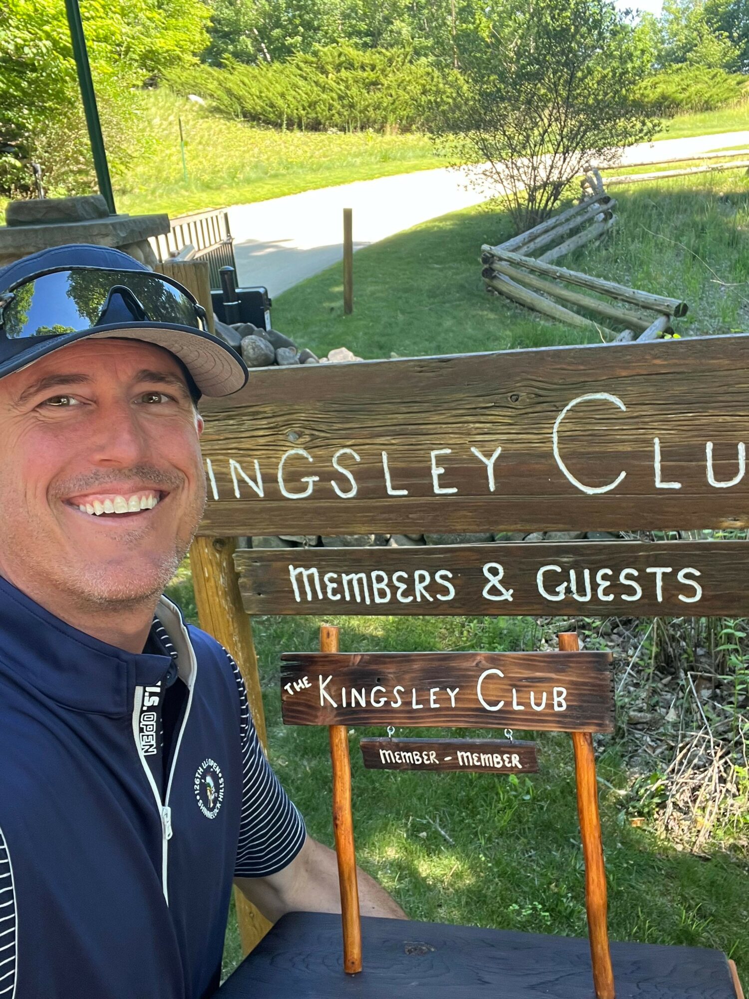 Smiling man wearing a hat and jacket outdoors near a wooden sign with trees and grass in the background.