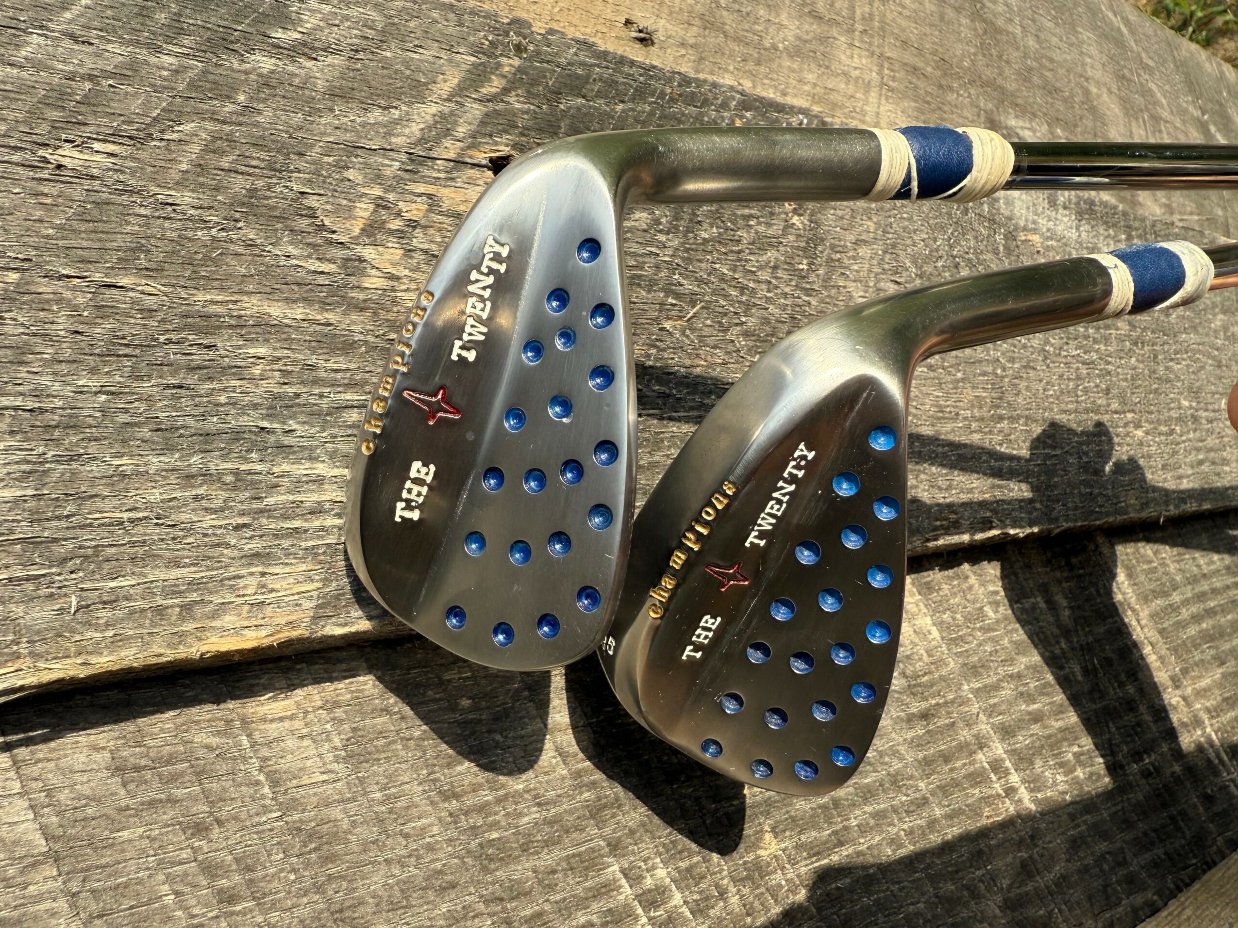 Two golf clubs with black and blue grips resting on a wooden surface, with shadows cast nearby.