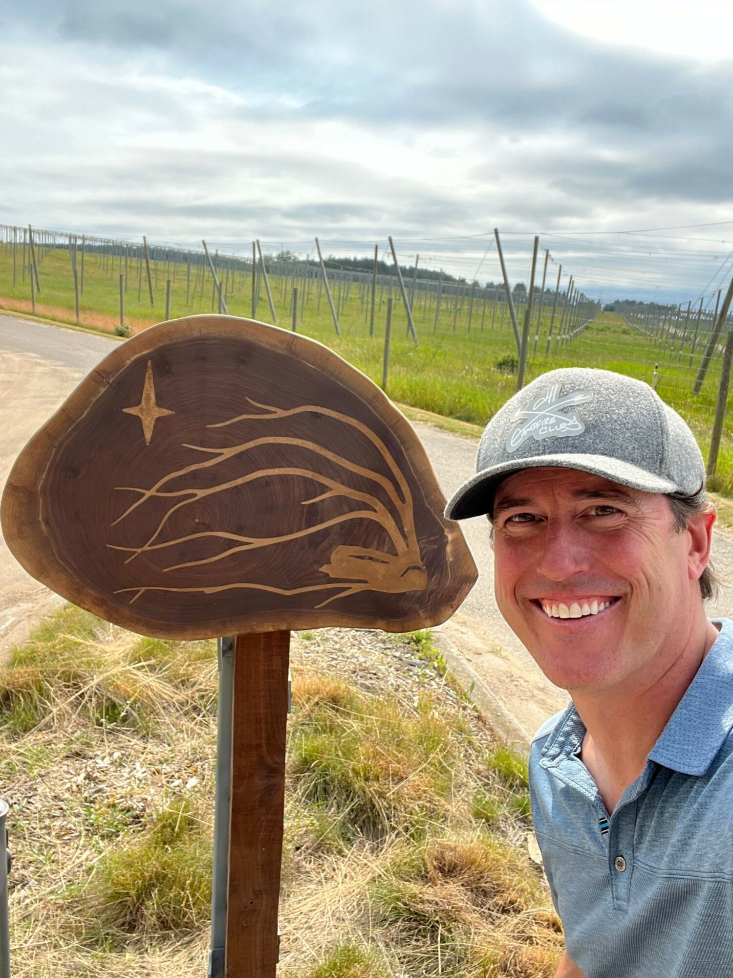 Person smiling outdoors near a wooden sign with a star, wearing a hat and blue shirt.