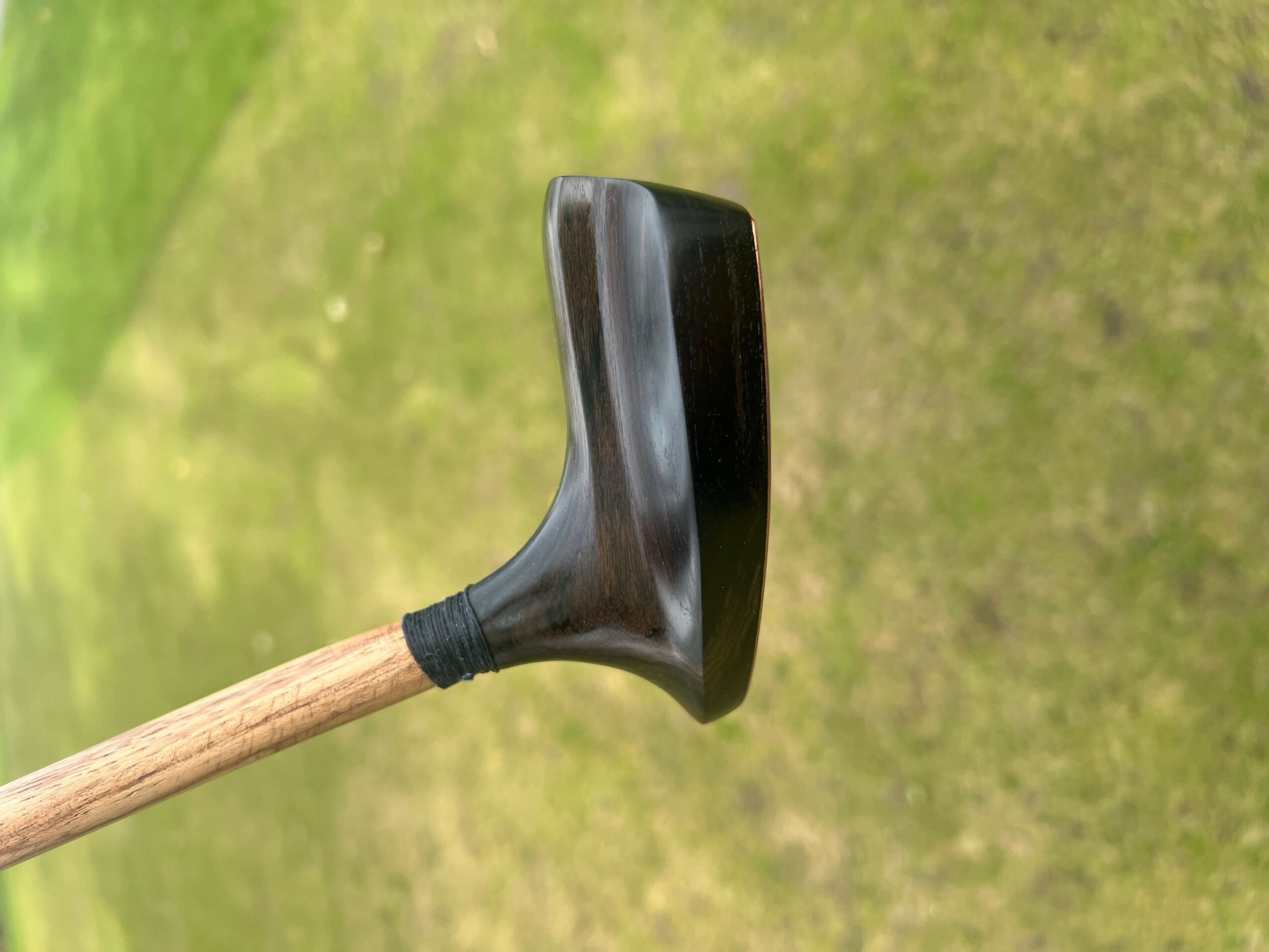 A black rubber mallet head attached to a wooden handle, positioned vertically against a grassy background.