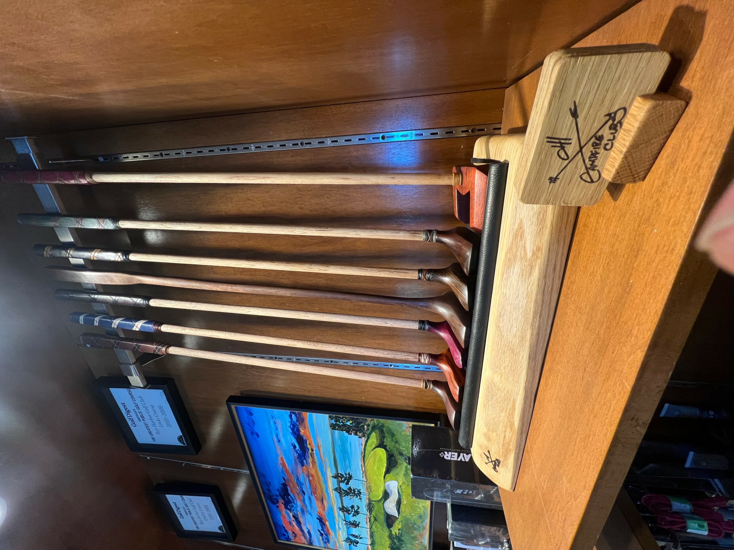 Golf clubs are stored vertically against a wooden wall, with a small wooden table holding a golf tee and a smartphone.