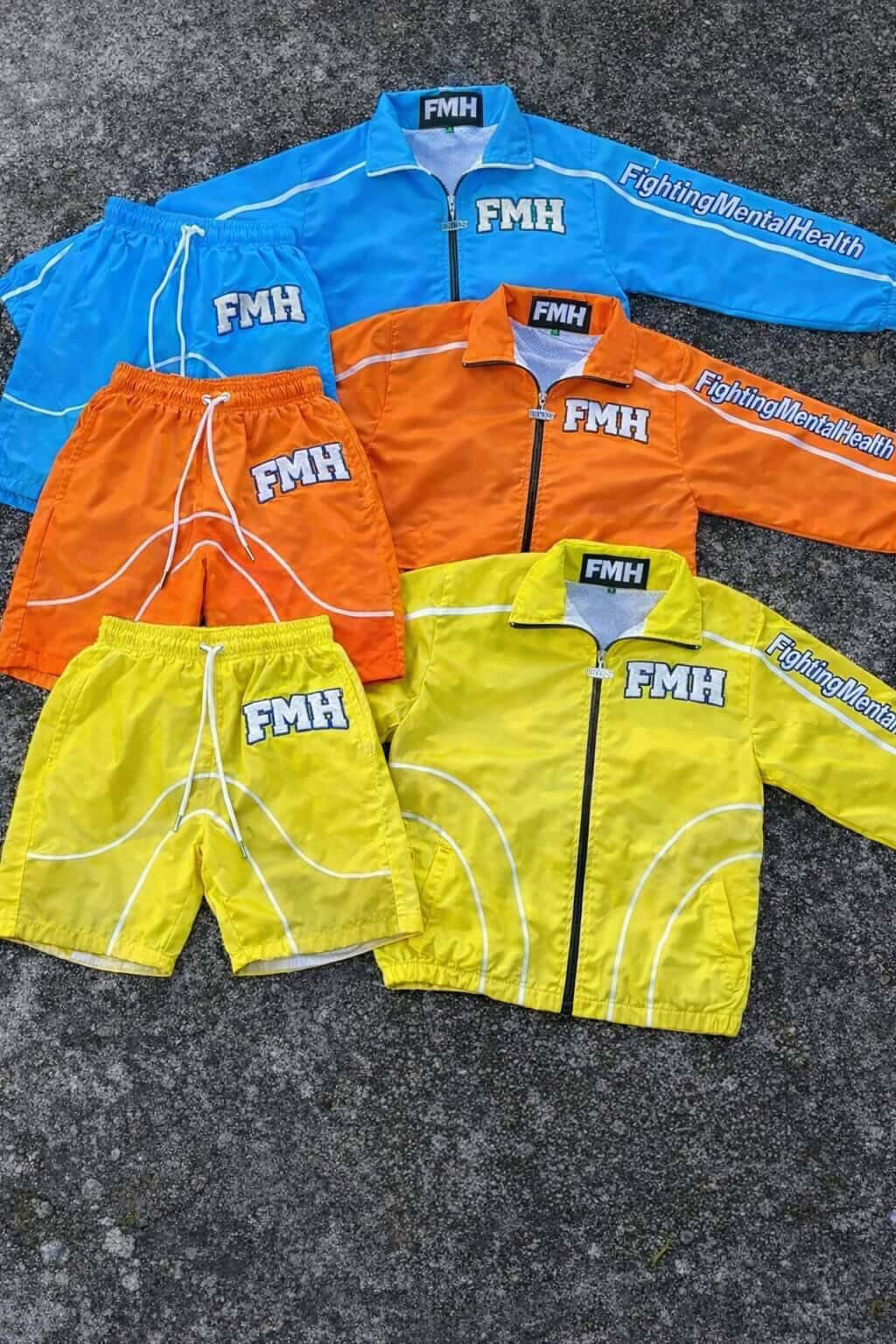 Four pairs of colorful shorts in blue, orange, yellow, and green with FMH logos, laid on a dark surface.