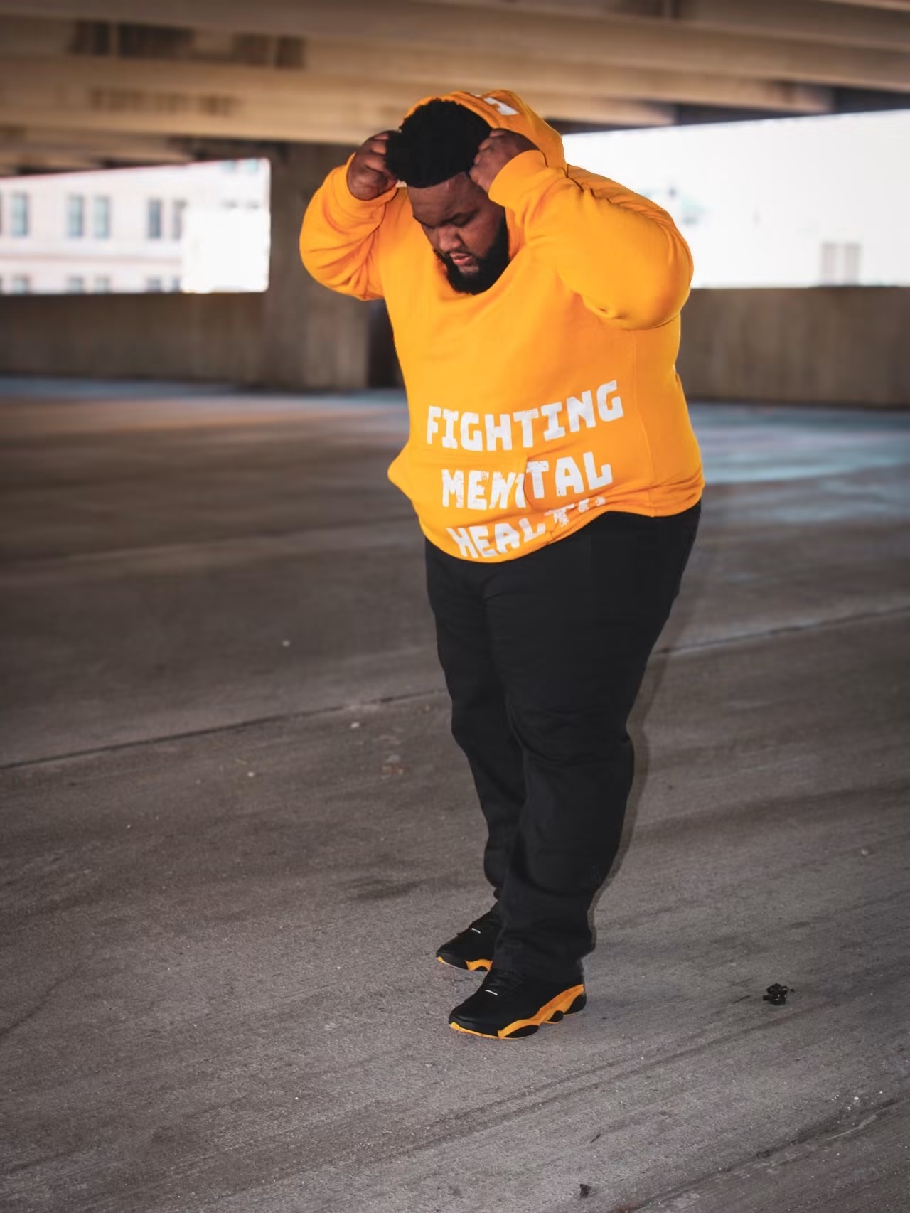 Person walking under a bridge wearing an orange hoodie with text, black pants, and sneakers, holding head with both hands.