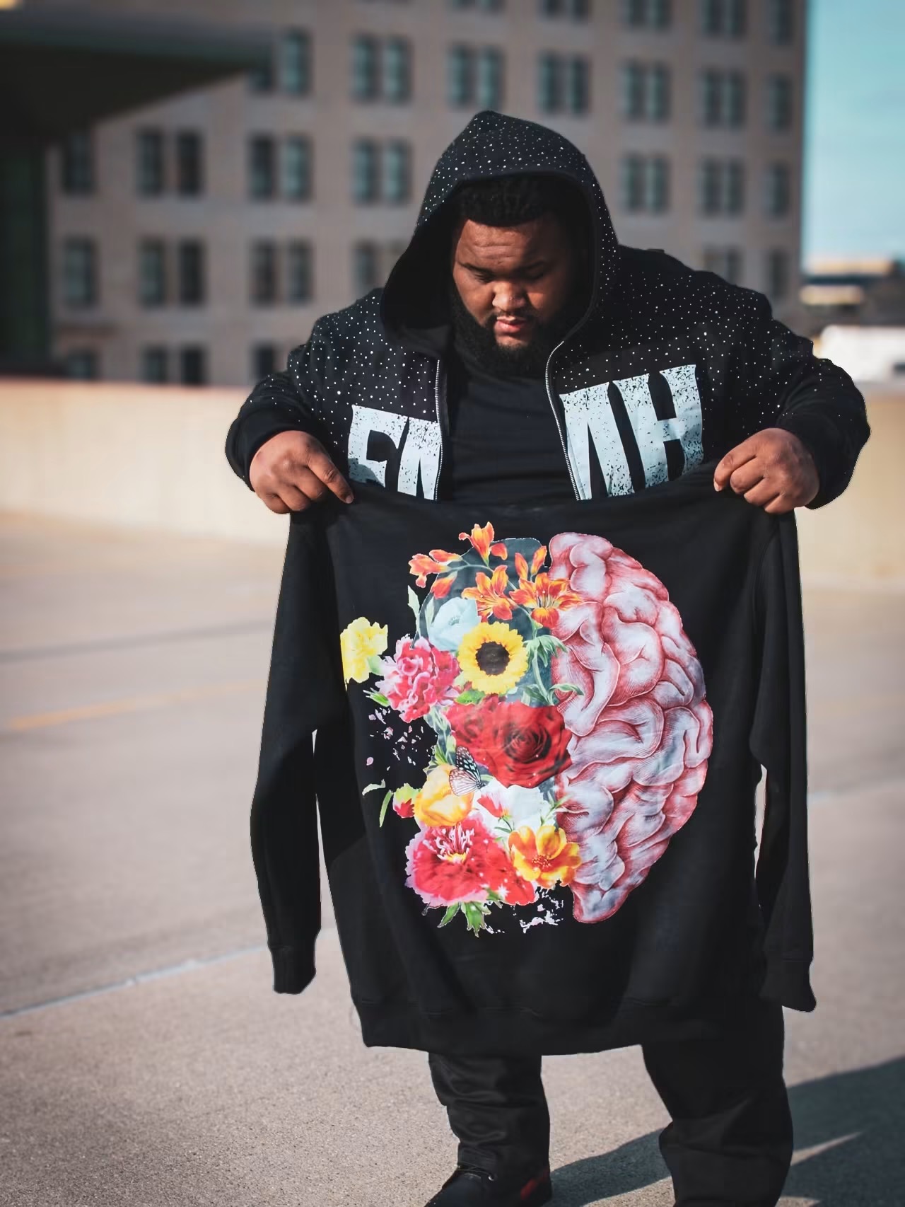Person holding a black cloth with a colorful brain and flower design, wearing a black hoodie, on a rooftop with city buildings.
