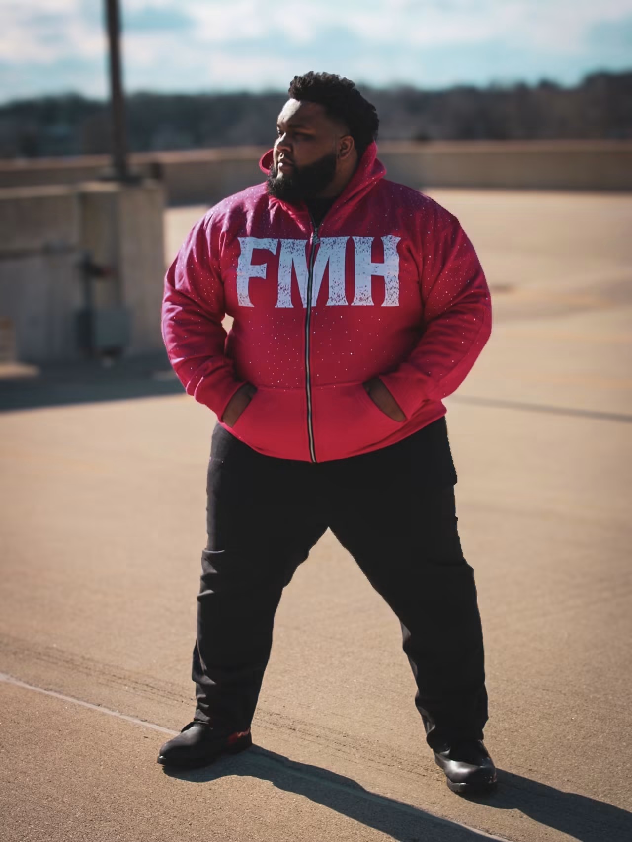 Man standing outdoors wearing a red hoodie with blue letters, hands in pockets, looking to the side.