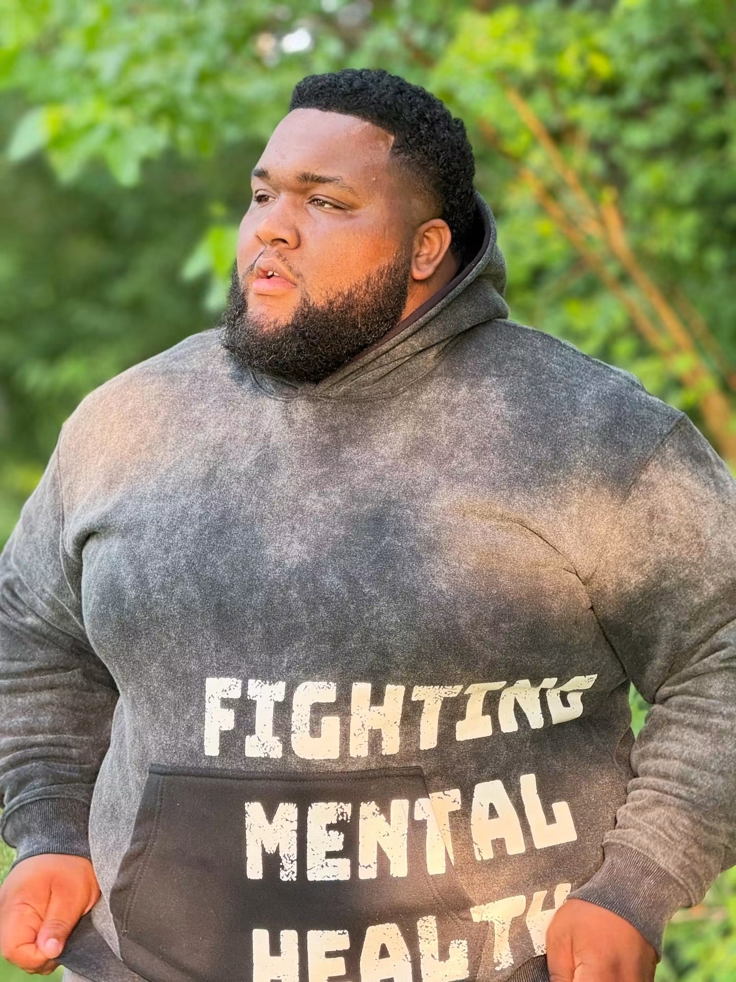 Man with beard and short curly hair wearing a gray hoodie outdoors, trees in background.