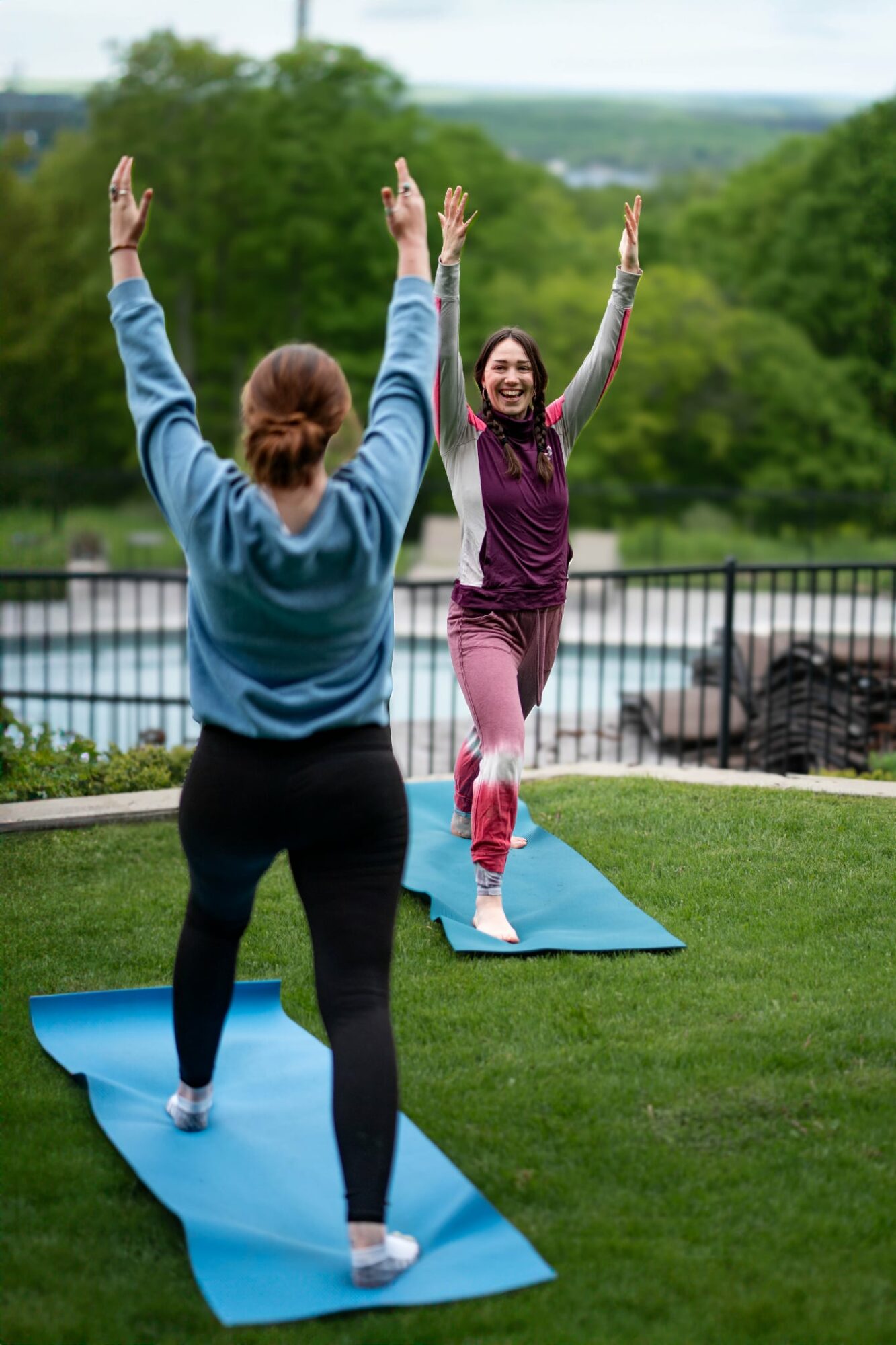Two women practicing yoga outdoors on mats, raising arms, with trees and a fence in the background.