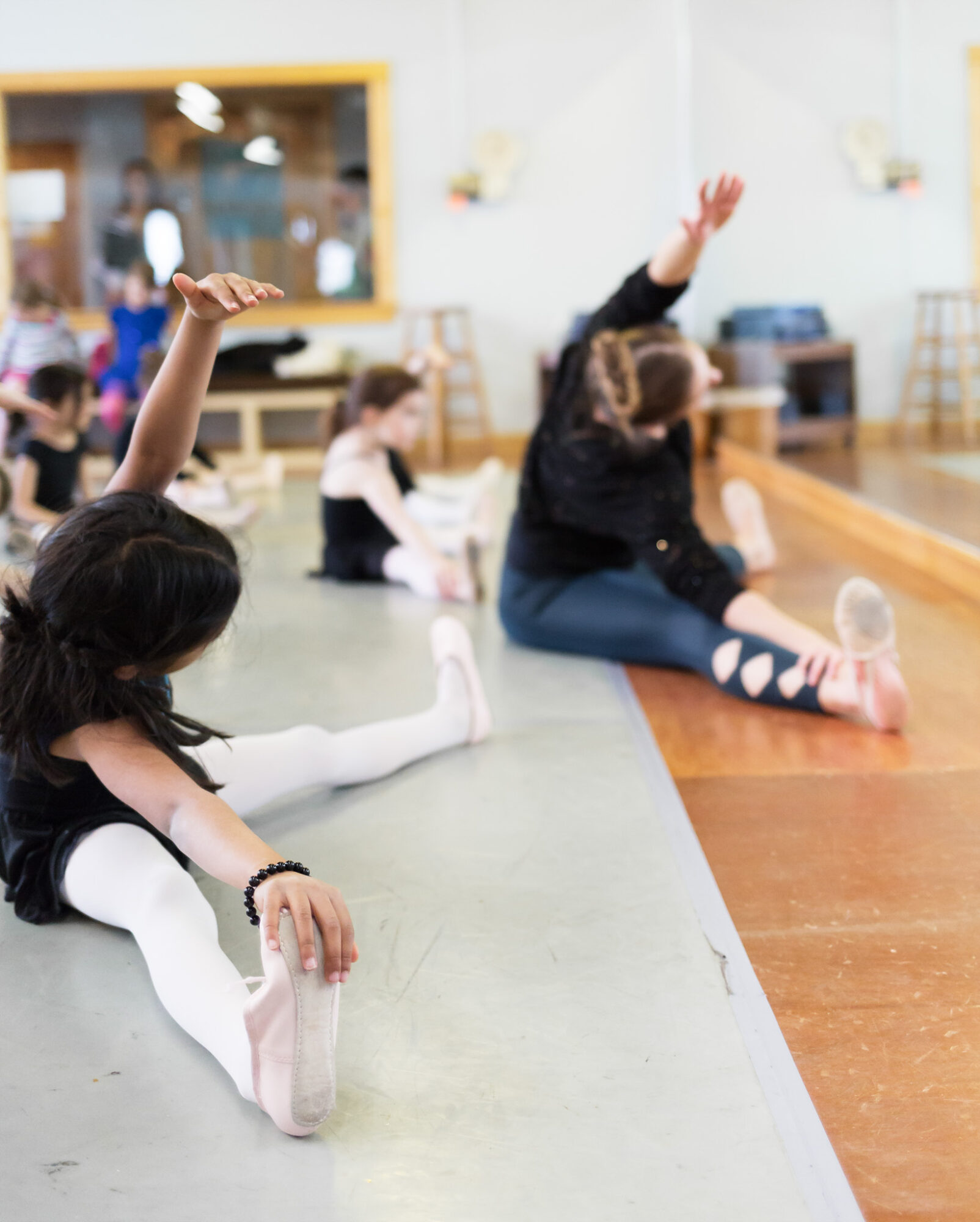 Children sitting on the floor in a dance studio, raising their hands during a class, with a mirror and wooden floor visible.