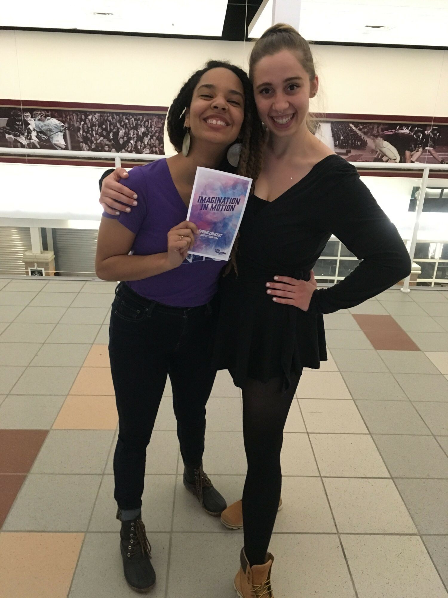 Two women standing in a shopping mall, one holding a colorful brochure, smiling at the camera.