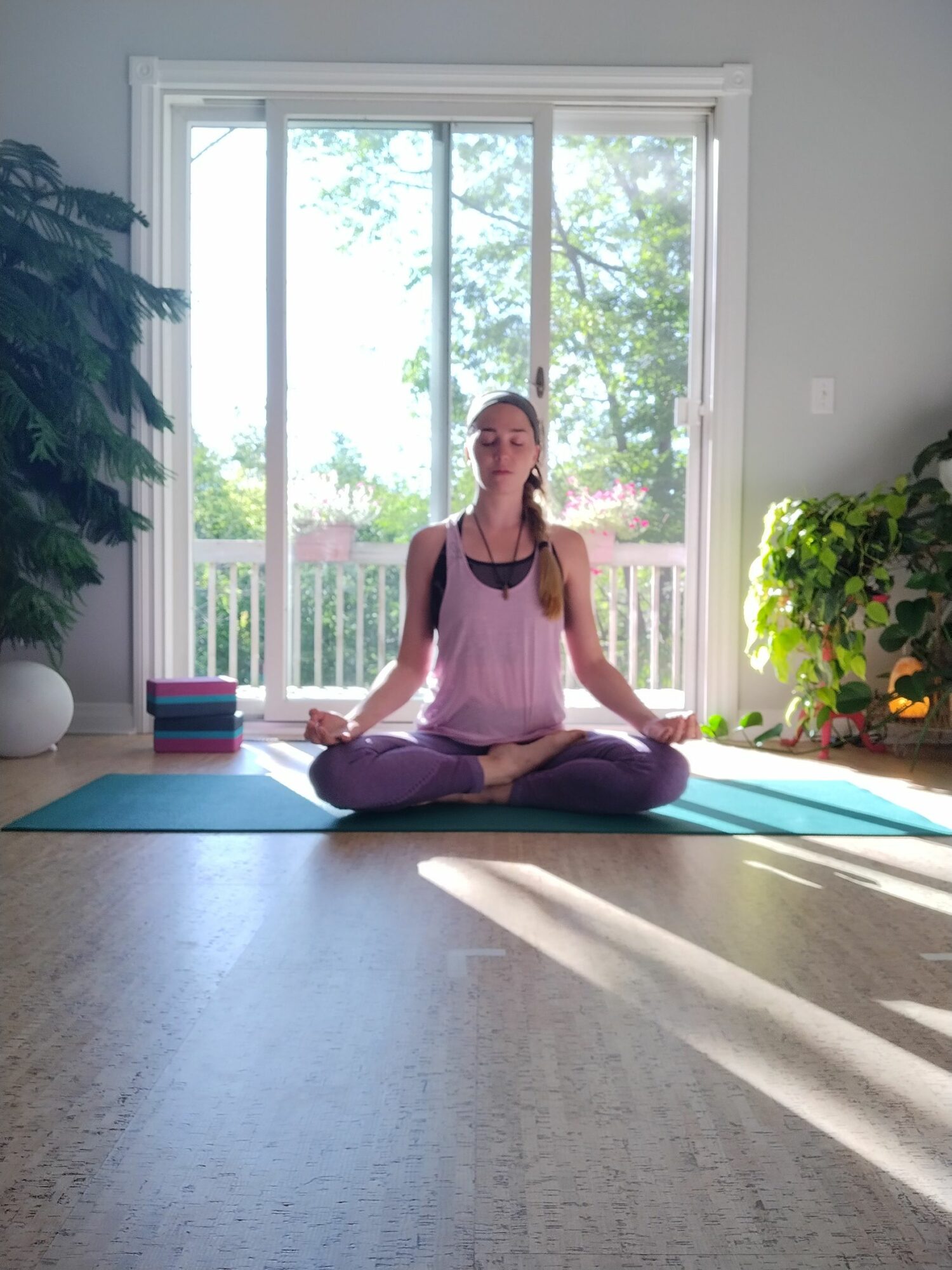 Woman practicing yoga on mat in bright room with large window and greenery outside.