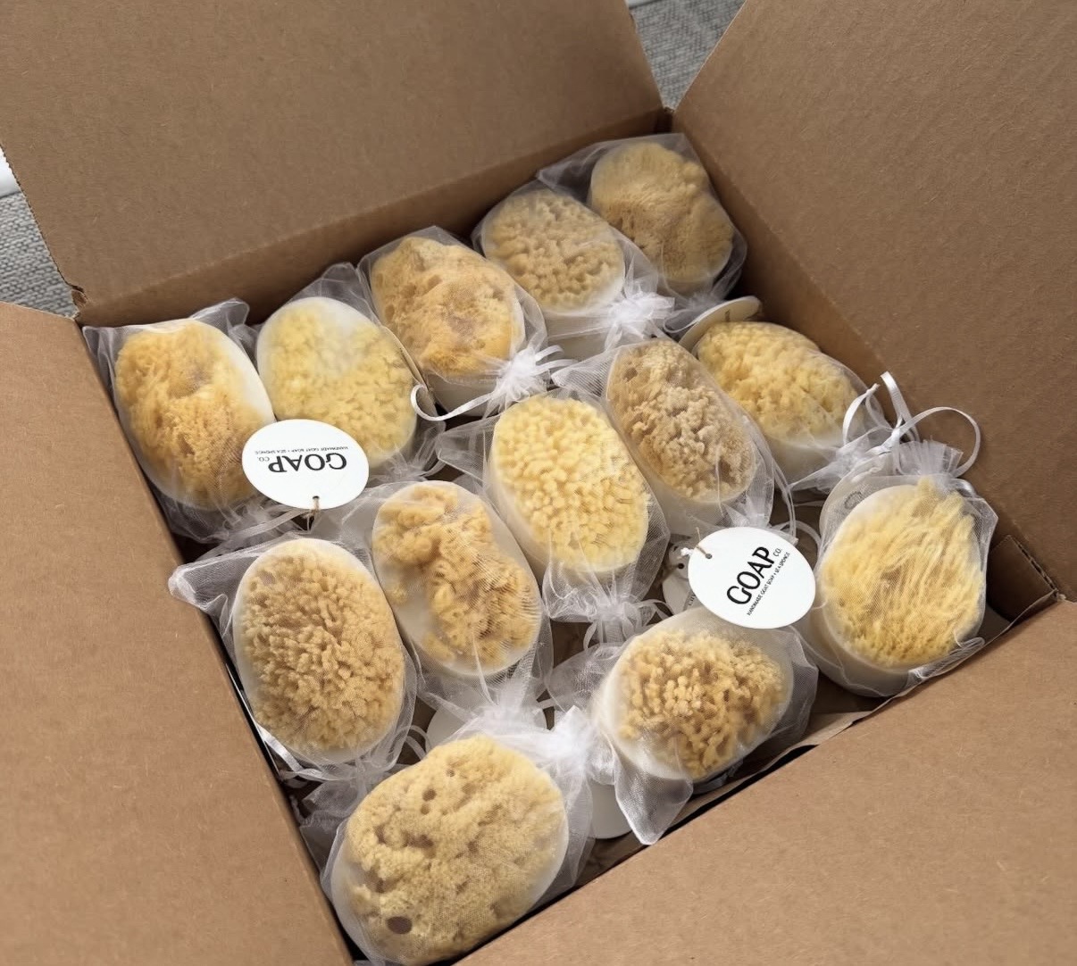 Open cardboard box containing multiple individually wrapped yellow sponge-like objects, some with tags labeled 'COA' and 'COV'.