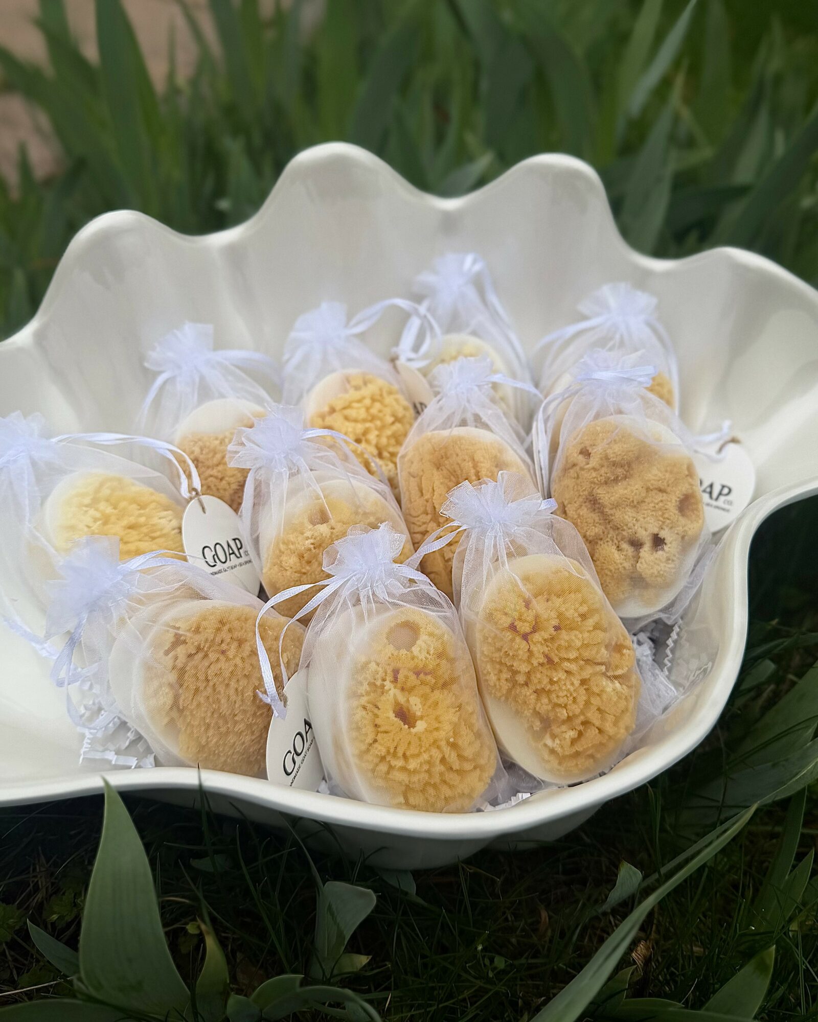 Cookies wrapped in white organza bags arranged in a white scalloped bowl outdoors with green grass and plants.