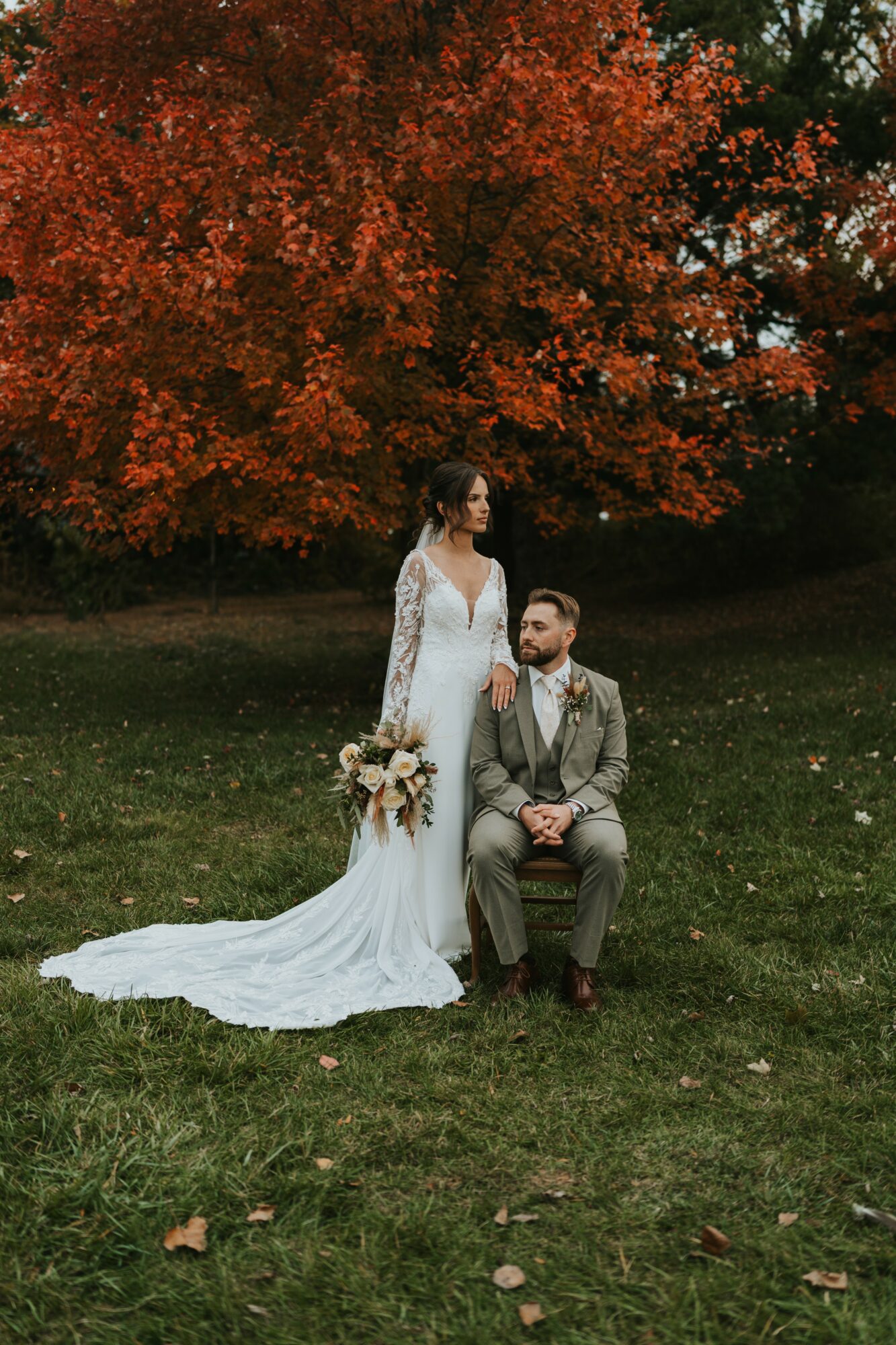 Bride and groom outdoors with a large autumn tree in background, bride standing, groom sitting on grass, bride holding bouquet.